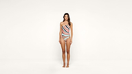 A woman stands barefoot against a plain white background, wearing a one-shoulder swimsuit with pink, black, and light blue wavy patterns. She faces forward with her arms by her sides.