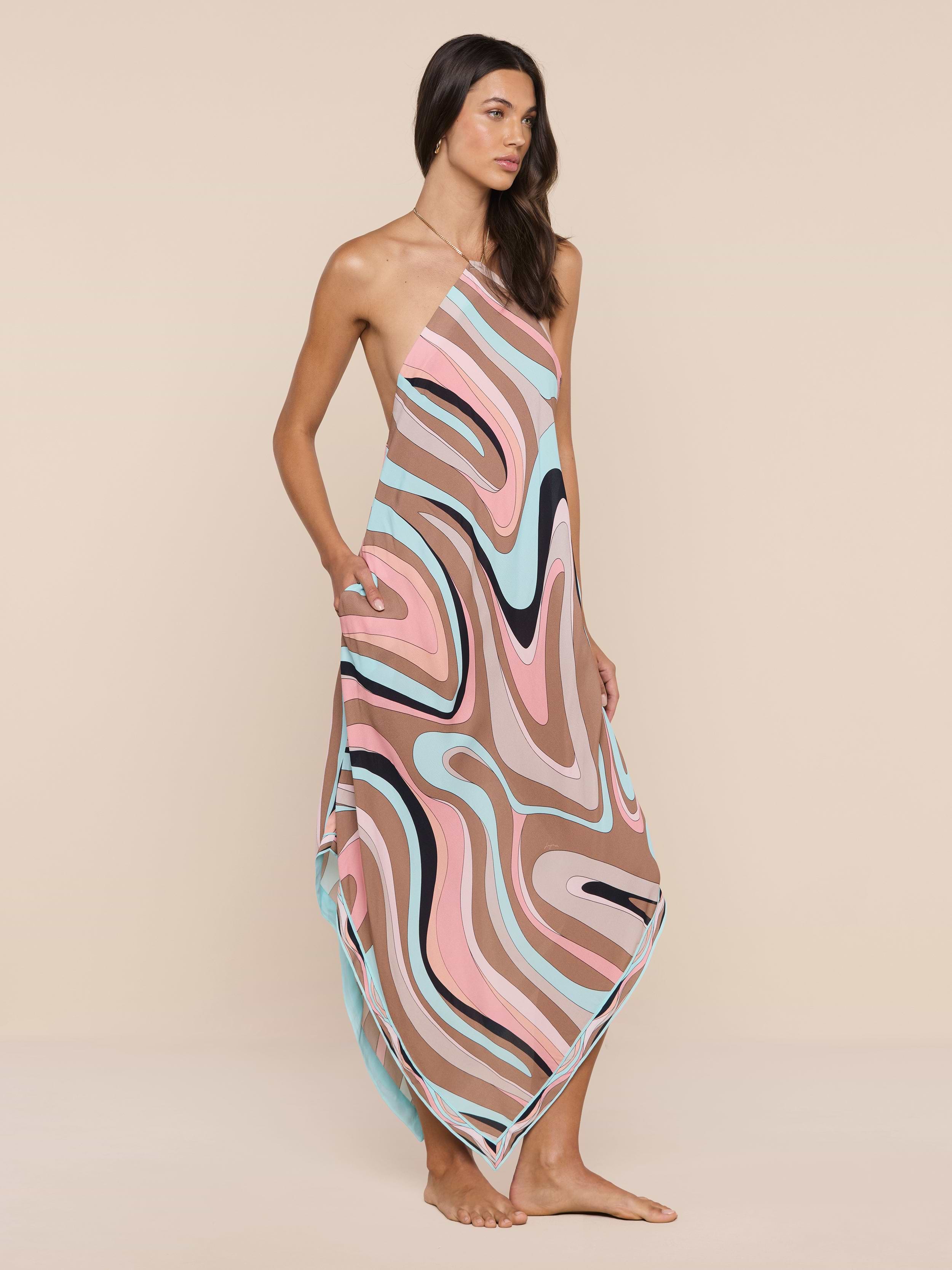Eine Frau steht barfuß in dem Elise Cover-Up Dress, einem wasserabweisenden Ein-Schulter-Krepp-Kleid mit einem abstrakten Wirbeldruck in Rosa-, Braun-, Blau- und Schwarztönen vor einem schlichten beigen Hintergrund.