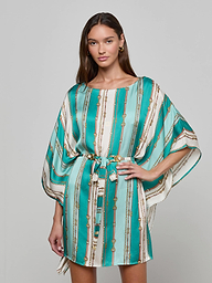 A woman wearing the Alia Cover-Up Tunic in a short, turquoise and white pattern with gold accents, wide sleeves, and a fabric belt at the waist stands against a plain background.