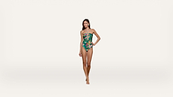 Woman standing against a white background, wearing a green one-piece swimsuit with a colorful floral pattern. She has one hand on her hip and is barefoot.