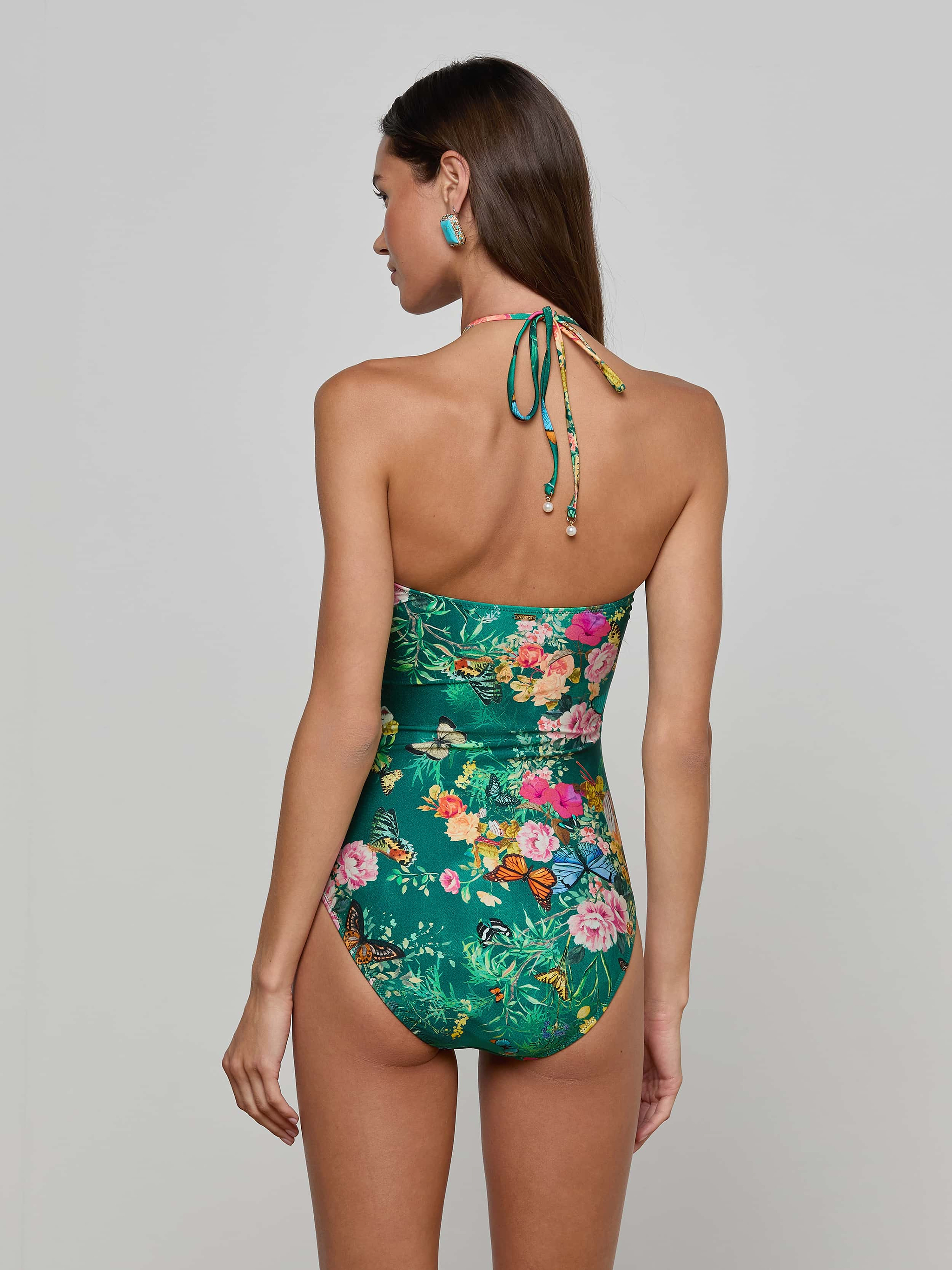 A woman stands with her back to the camera, wearing the Shiva One-Piece Swimsuit—a green butterfly floral print halter style—paired with her hair down and colorful earrings, set against a plain light background.
