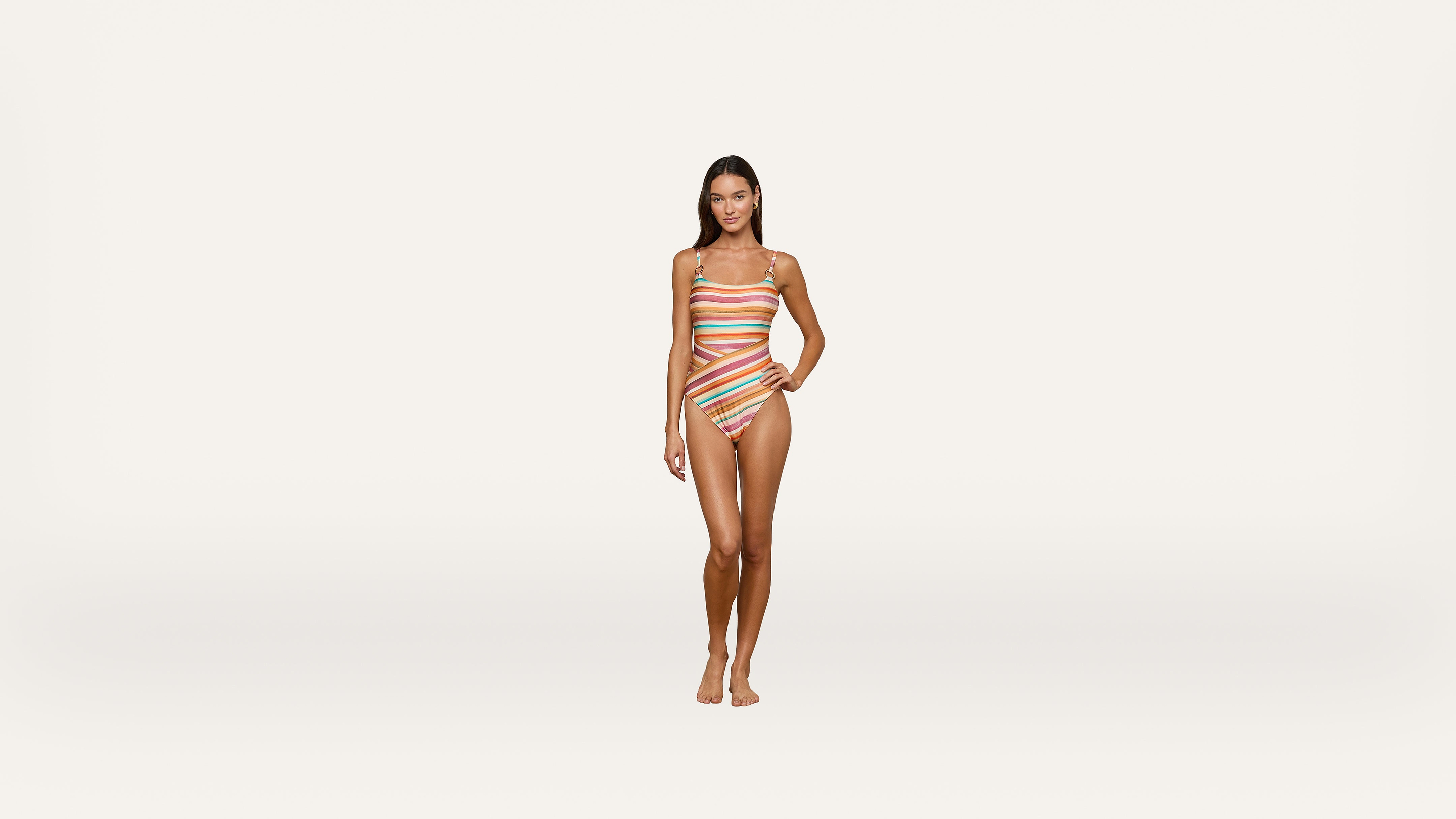 A woman stands facing forward, wearing a colorful, striped one-piece swimsuit with cutouts. She stands barefoot against a plain white background.