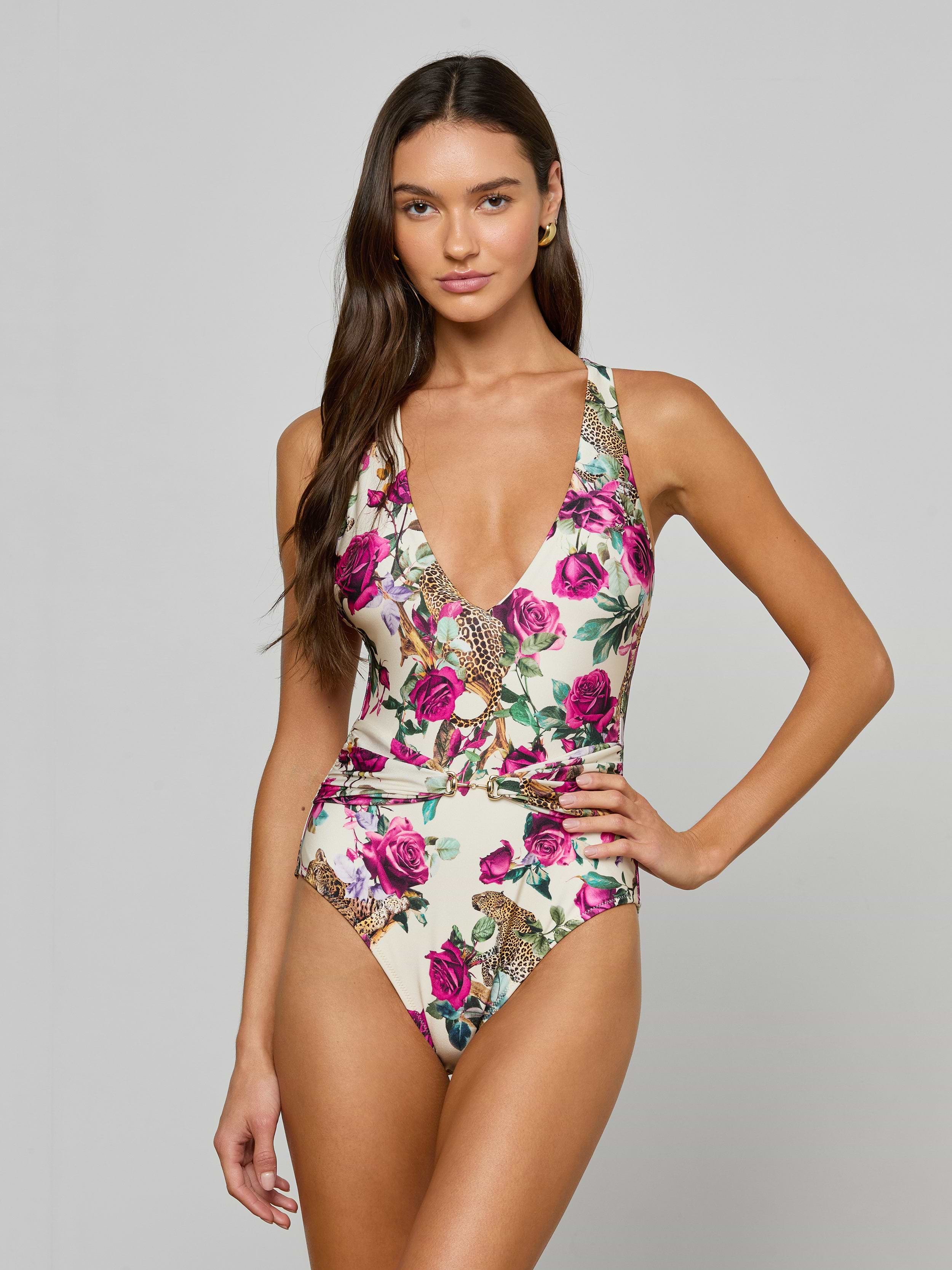 A woman wearing the Lisa Plunge One-Piece Swimsuit with a floral print and a deep V-neckline stands against a plain light background, one hand on her hip, looking at the camera.