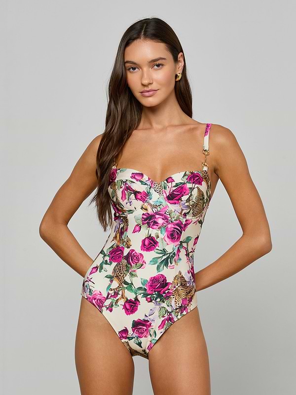 A woman stands against a plain light background, looking at the camera with her hands on her hips, wearing the Amie Bandeau One-Piece Swimsuit—a chic white one-piece featuring a pink floral and leopard print design that combines style and comfort.