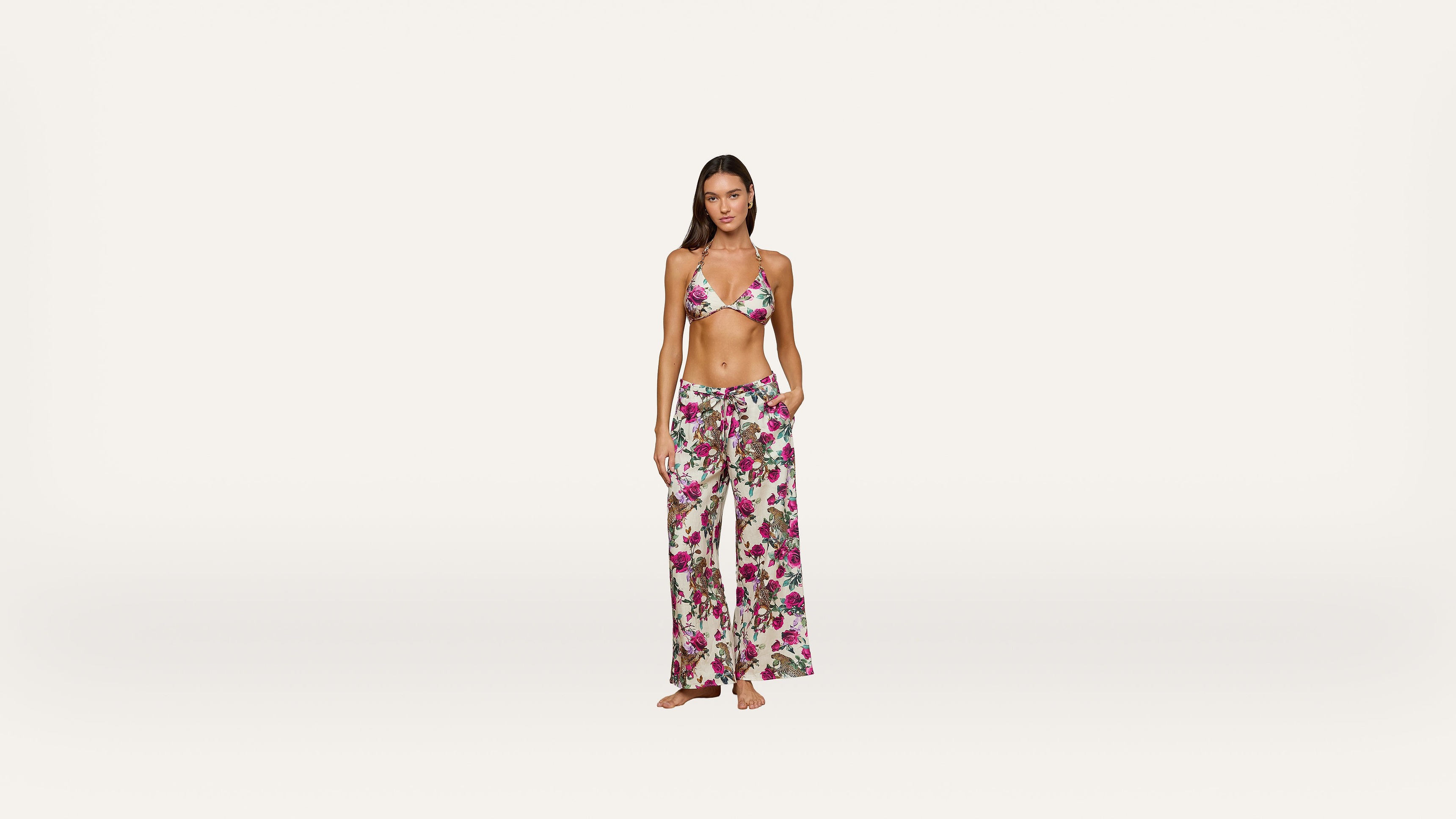 Woman standing barefoot against a plain background, wearing a floral bikini top and matching wide-leg pants with pockets. She has long hair and a relaxed expression, with one hand in her pocket.