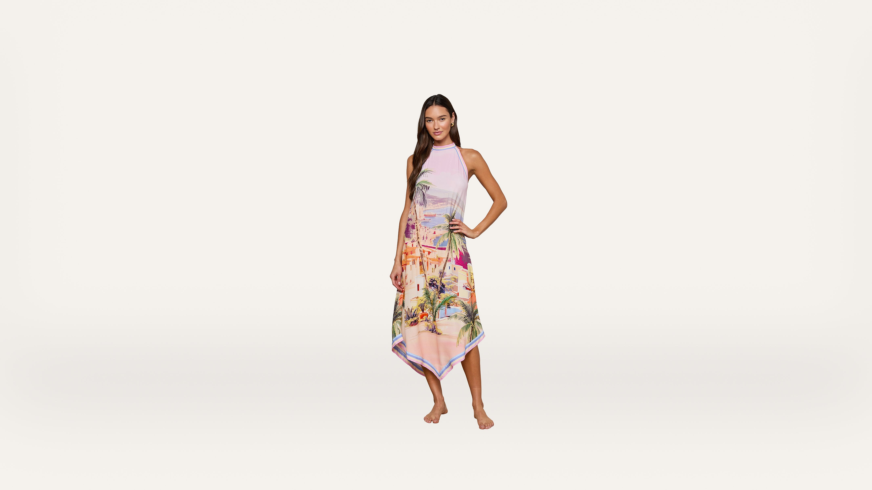 A woman stands barefoot, wearing a colorful, printed halter-neck dress with a handkerchief hem, against a plain white background.