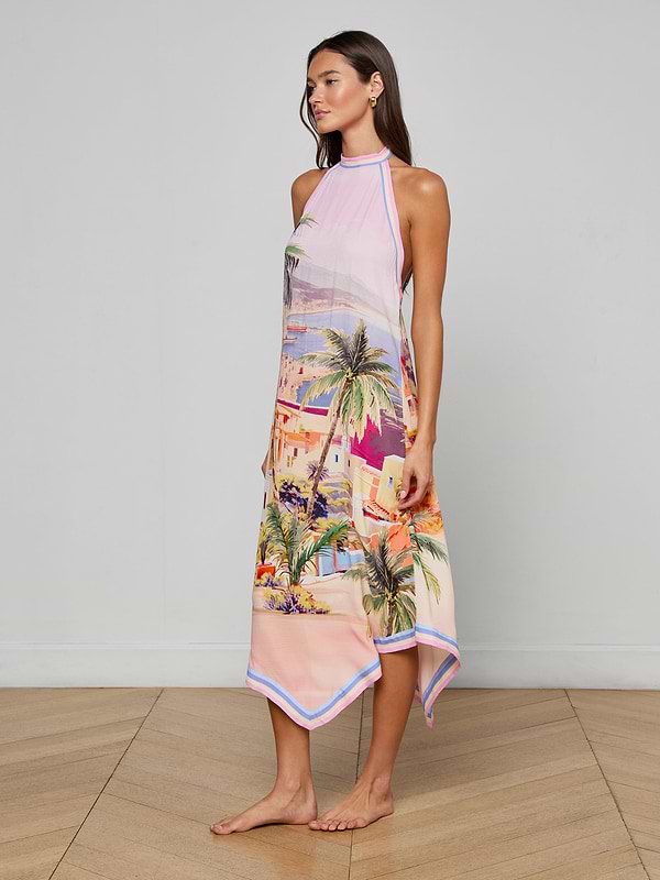 A woman stands barefoot wearing the Elise Cover-Up Dress, a water-safe crepe halter midi dress adorned with a vibrant scenic postcard print of palm trees and buildings, set against a plain white background.
