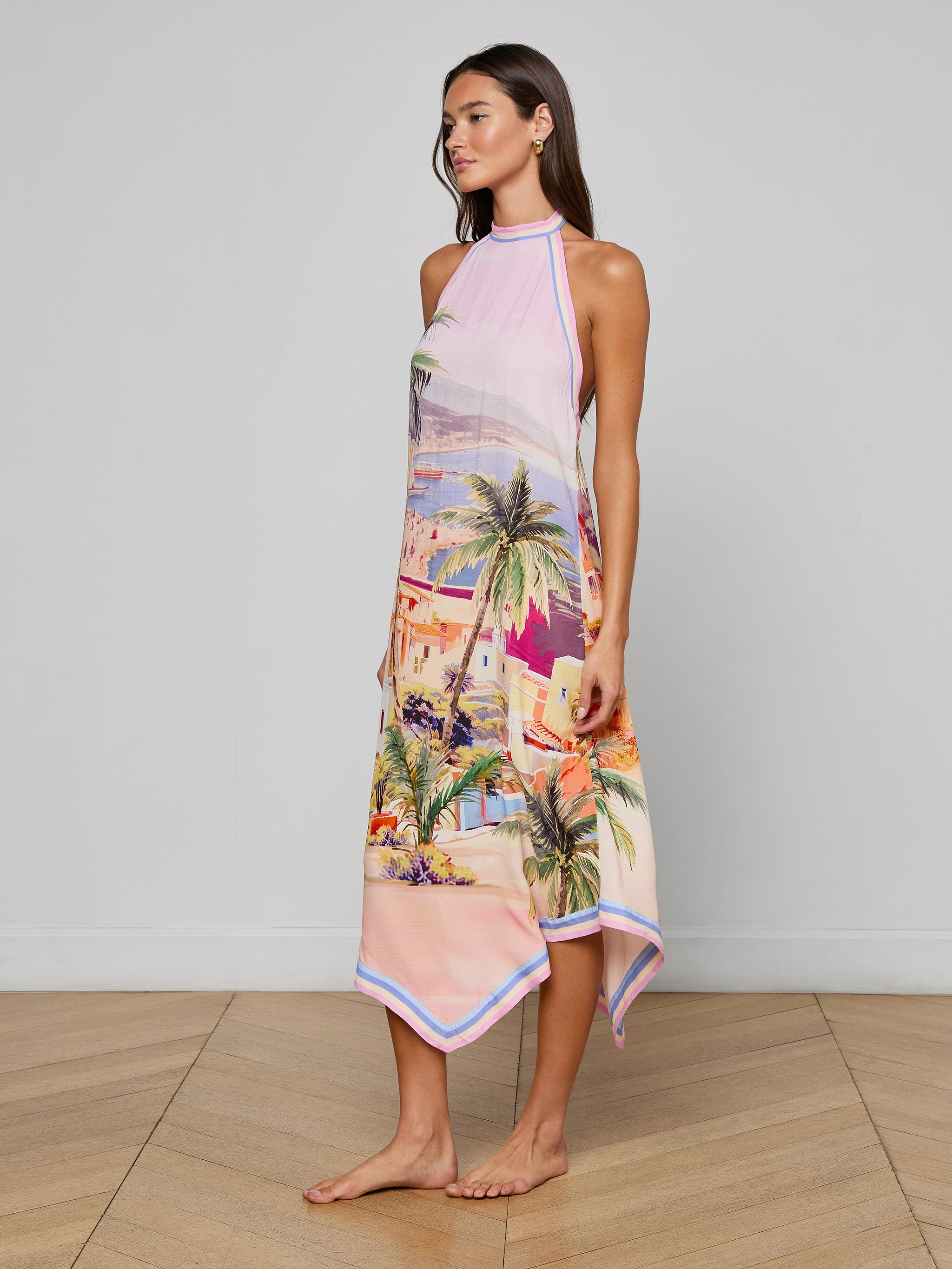 A woman stands barefoot wearing the Elise Cover-Up Dress, a water-safe crepe halter midi dress adorned with a vibrant scenic postcard print of palm trees and buildings, set against a plain white background.