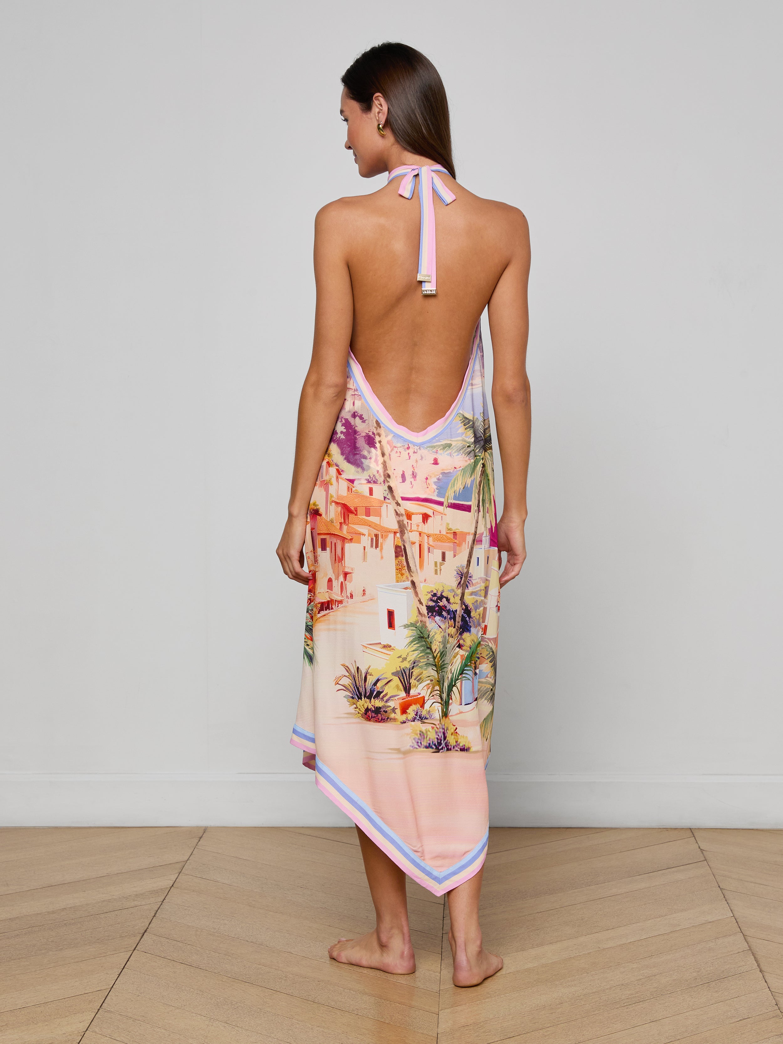 A woman stands barefoot, shown from the back, wearing the Elise Cover-Up Dress—a water-safe crepe halter midi dress with an open back and a colorful scenic postcard print depicting buildings and palm trees. The background is plain white.