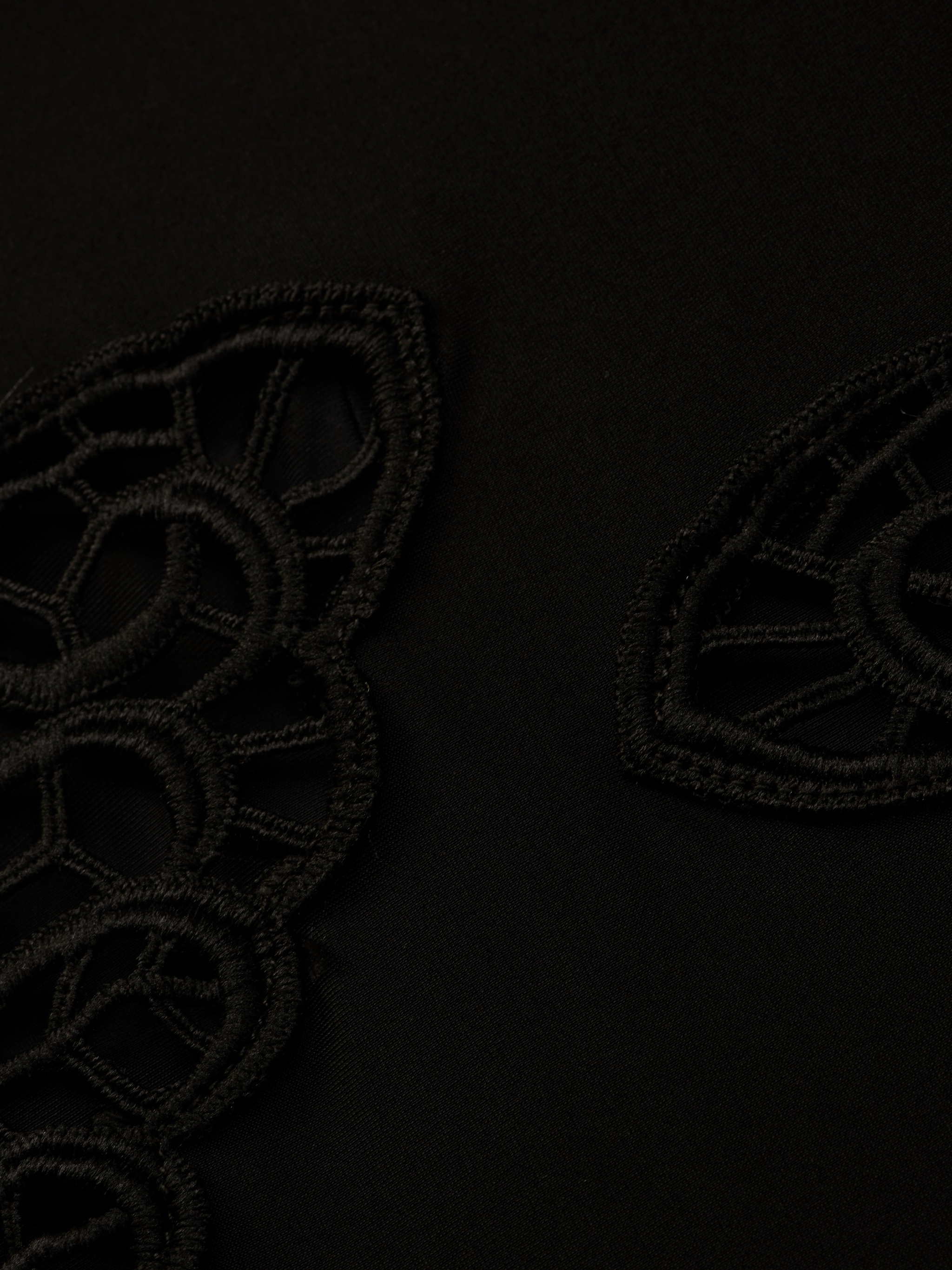 Close-up of the Remi One-Piece Swimsuit’s black fabric, featuring geometric, lace-like embroidered patterns reminiscent of poolside glamour. The textured embroidery creates a subtle contrast against the smooth background.