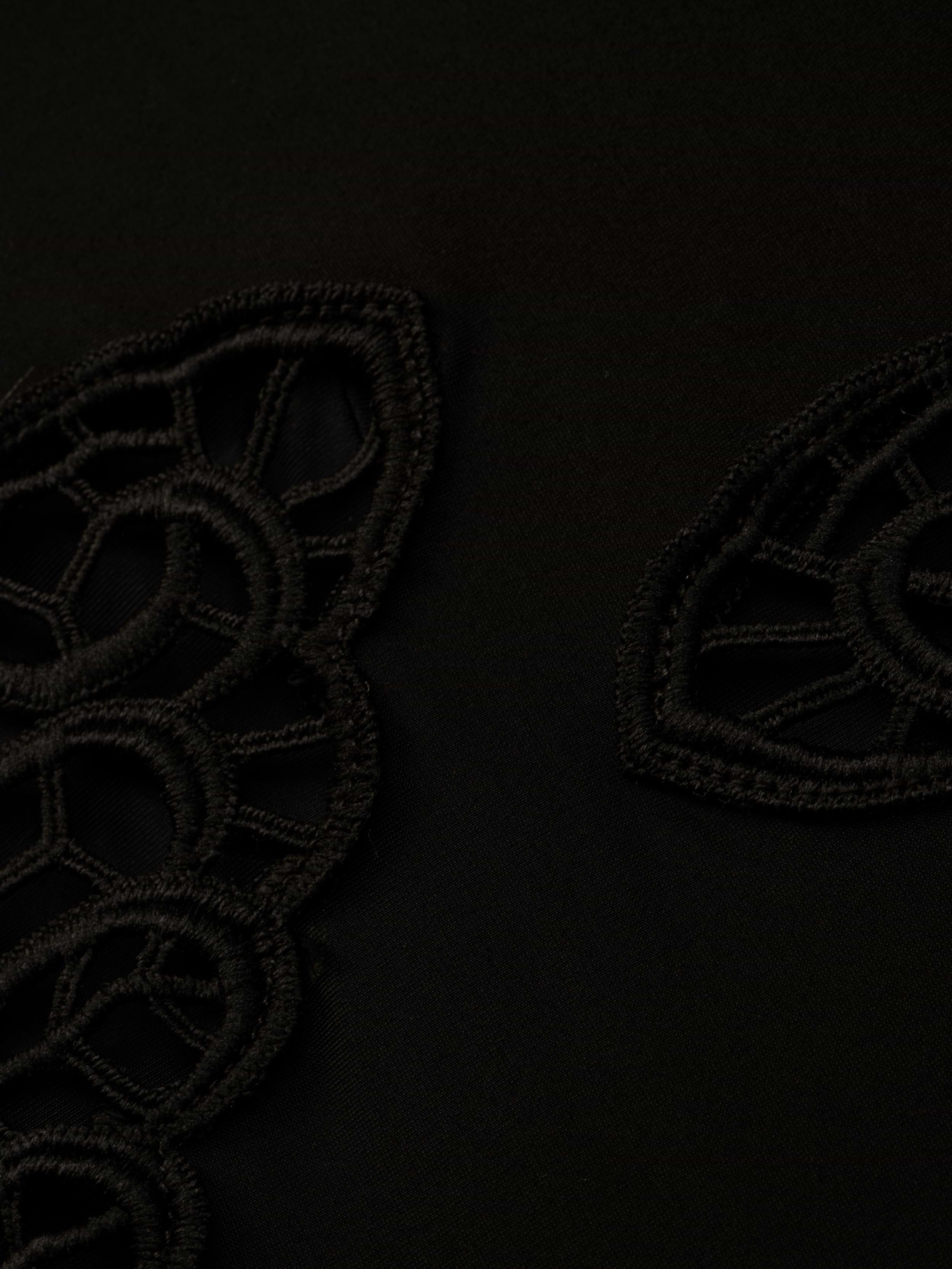 Close-up of the Remi One-Piece Swimsuit’s black fabric, featuring geometric, lace-like embroidered patterns reminiscent of poolside glamour. The textured embroidery creates a subtle contrast against the smooth background.