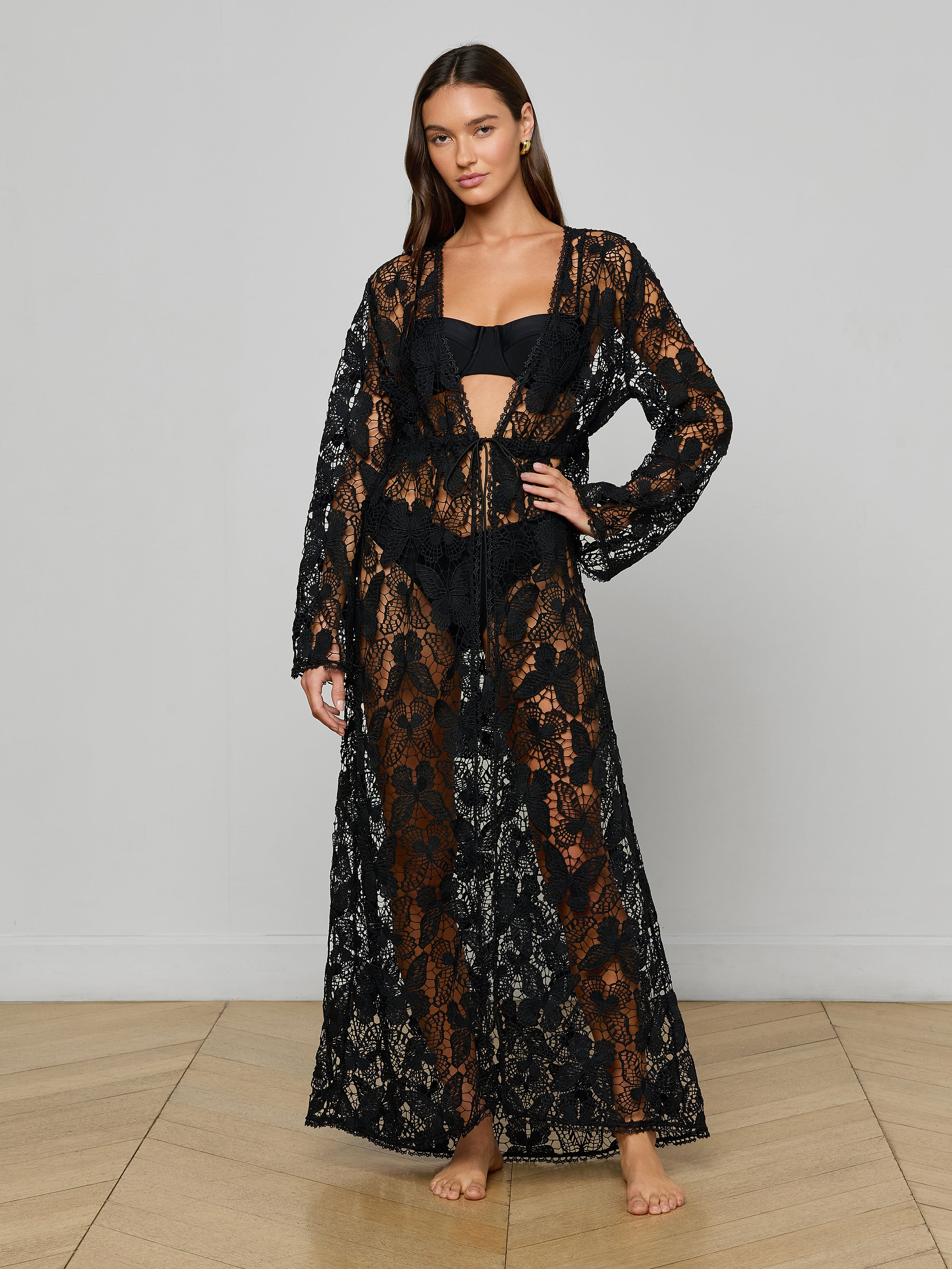 A woman stands barefoot against a plain white background, wearing the Janus Cover-Up Robe—a long-sleeve, sheer black lace bohemian layer—over a black bikini.
