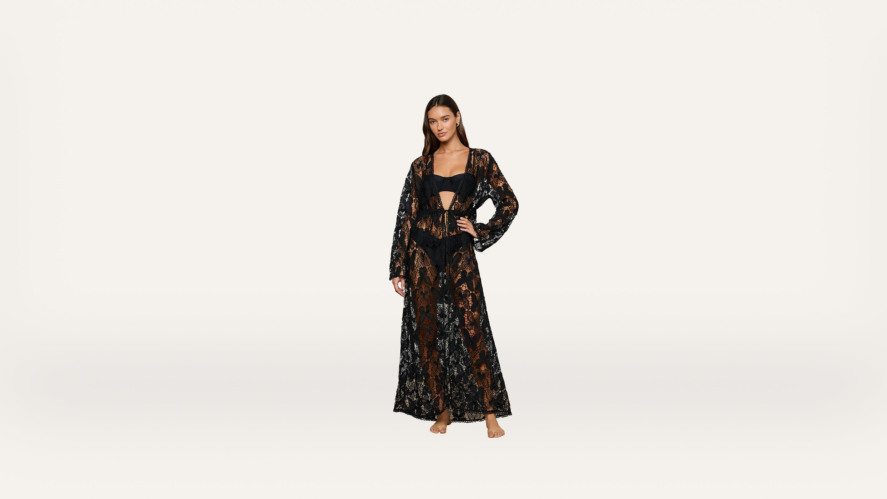 A woman stands barefoot, wearing a long, sheer black lace dress over a matching black bralette and high-waisted shorts, posing against a plain white background.