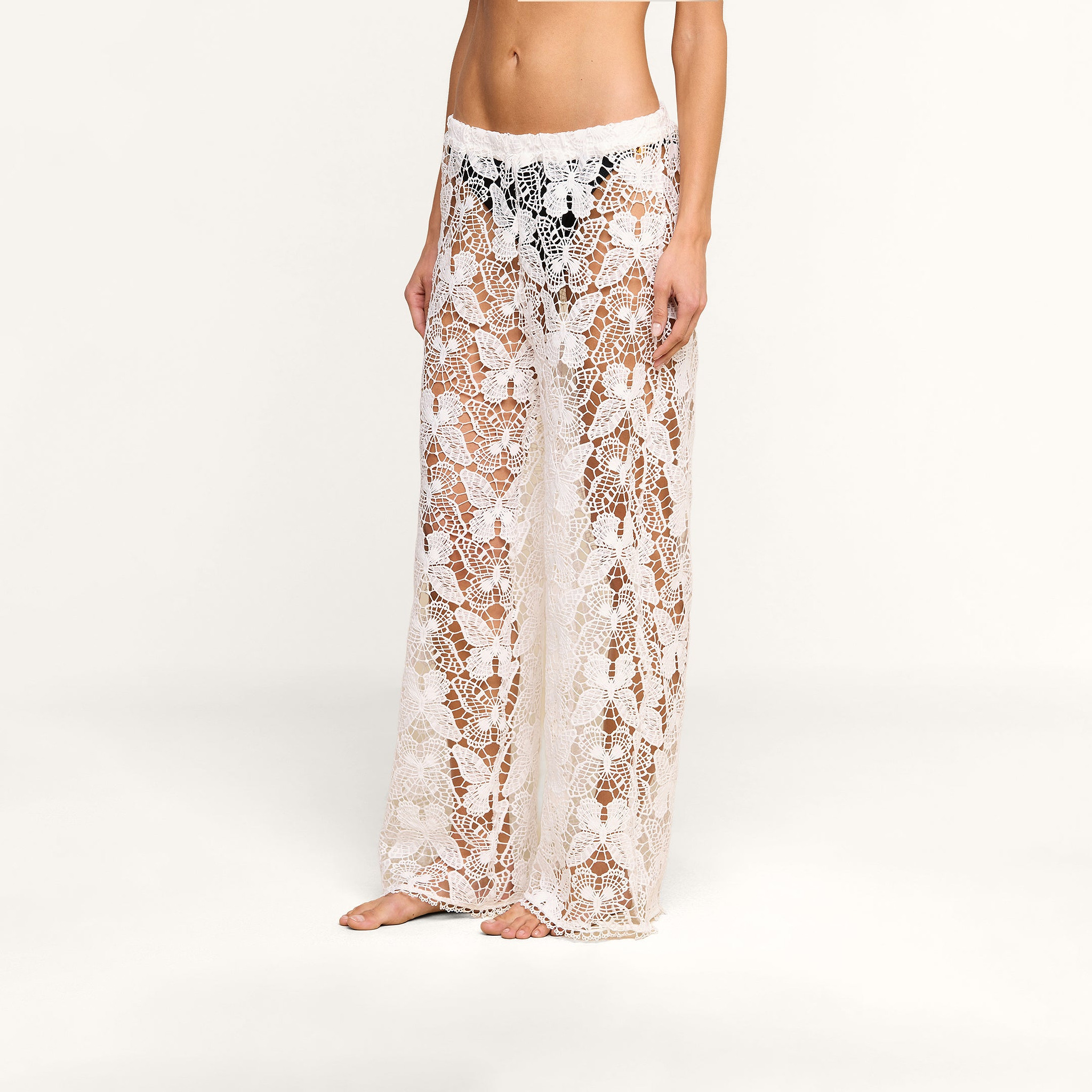 Una mujer luce los pantalones «Geraldine Cover-Up»: unos pantalones blancos de croché transparente con un delicado estampado de mariposas y flores que deja entrever sutilmente la ropa interior negra que lleva debajo. Ideales para llevar en un complejo turístico, la modelo posa descalza sobre un fondo beige liso.