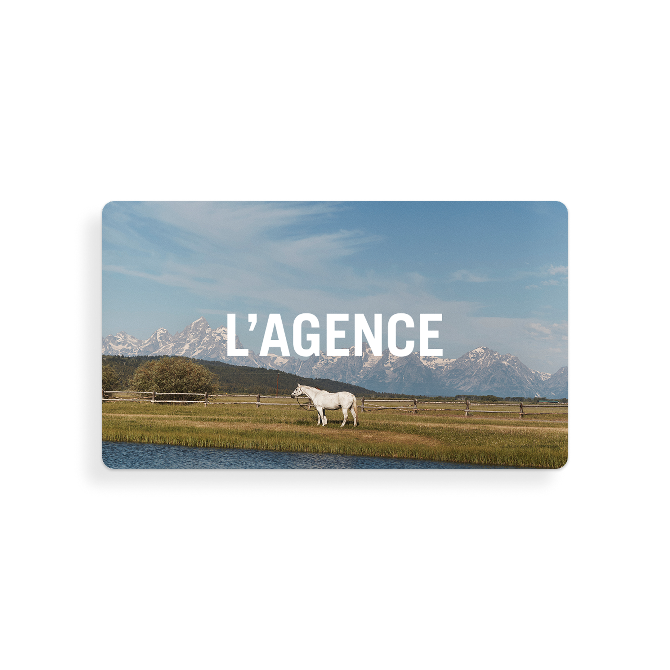 A white horse stands in a grassy field near water, set against a backdrop of mountains and blue sky. The image features the words “L’AGENCE Gift Card” in bold white text, evoking a sense of tranquility that mirrors the seamless experience of receiving thoughtful gift card redemption instructions.