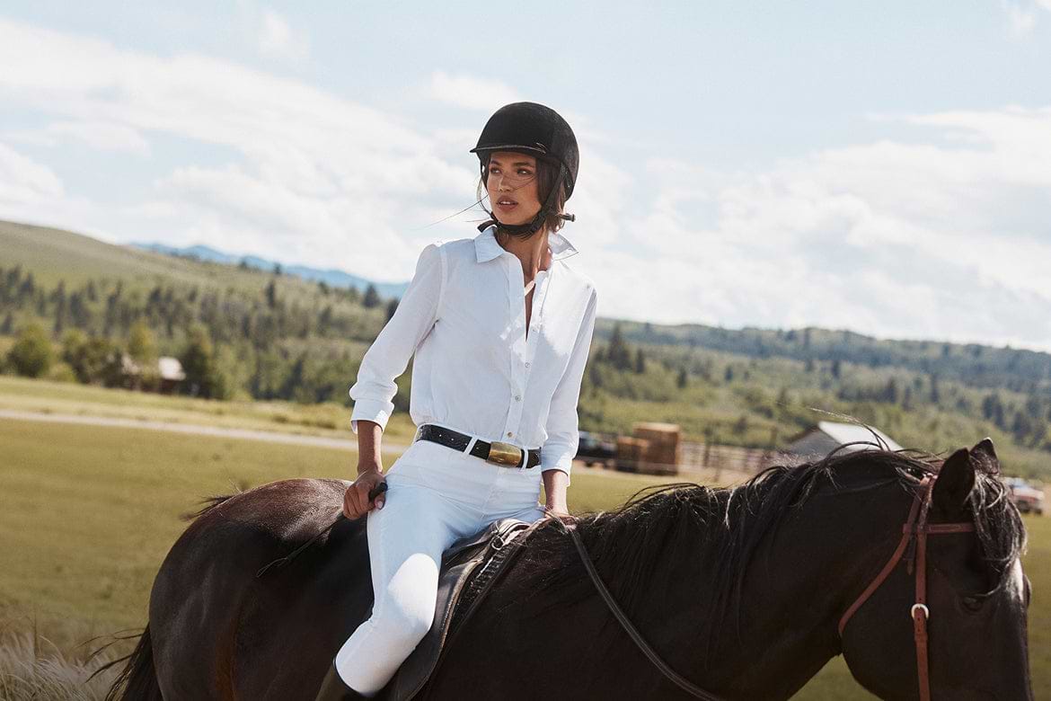 A person wearing a white shirt, white pants, and a black helmet rides a dark horse outdoors, surrounded by green fields and trees under a partly cloudy sky.