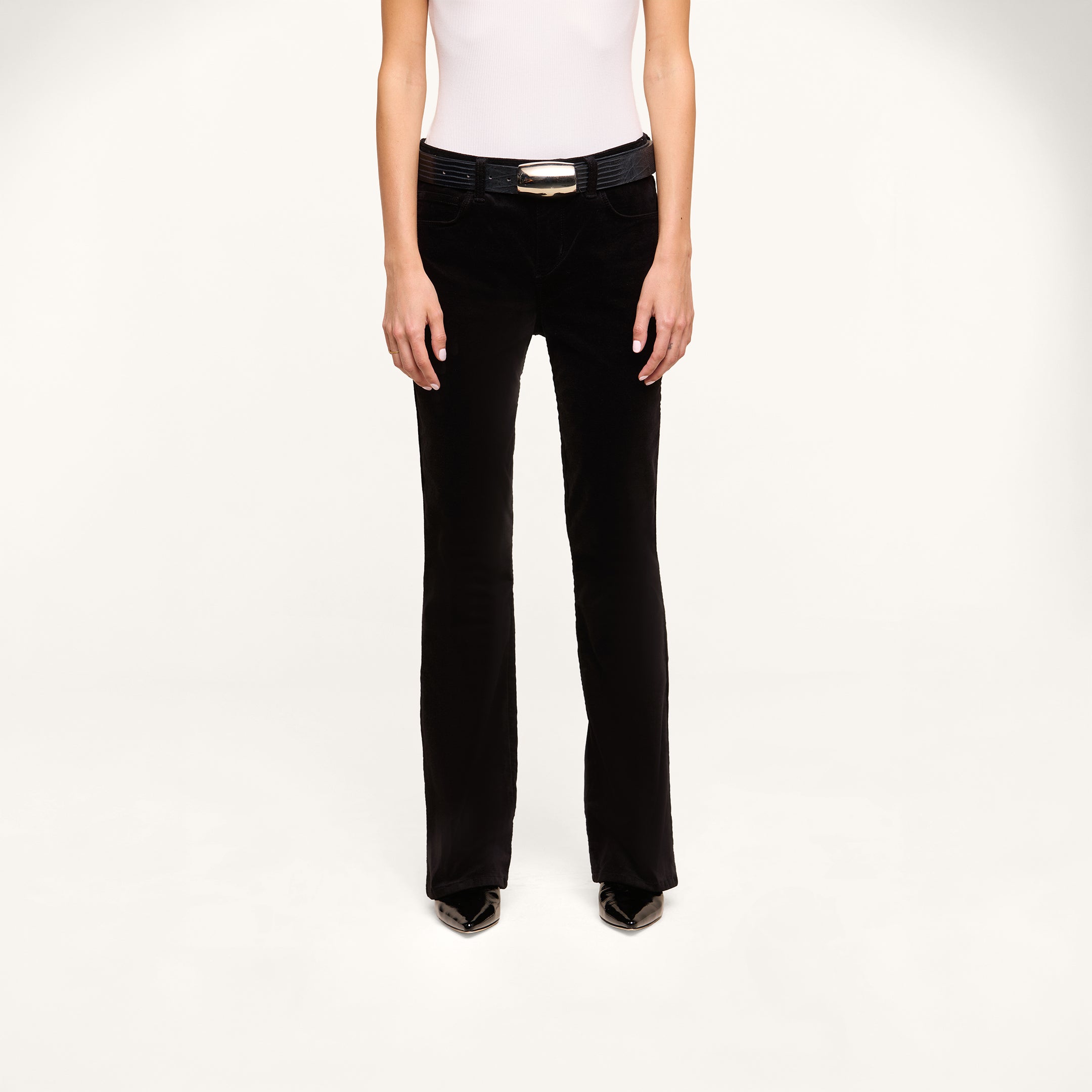 A person wearing a white sleeveless top, the Petite Stevie Velvet Jean with a belt, and black pointed shoes stands against a plain white background, facing forward.