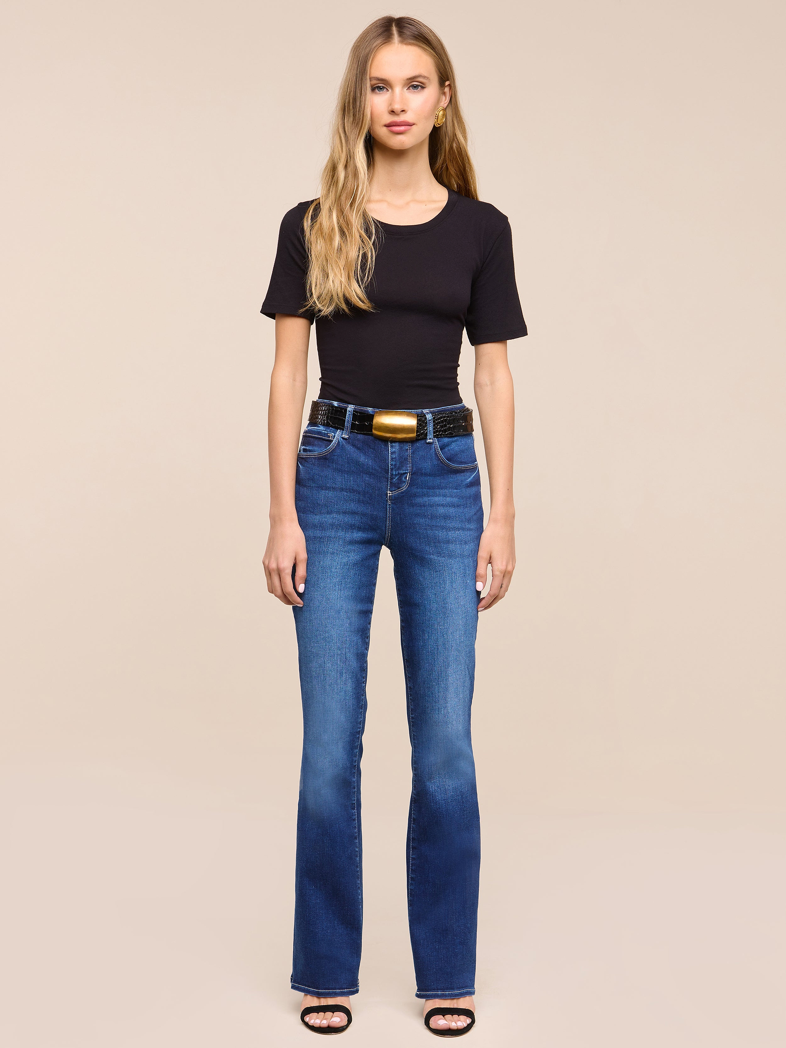 A woman with long blonde hair wears a black t-shirt, the Petite Selma Bootcut Jean in blue super stretch denim, a black belt with a gold buckle, and black sandals, standing against a plain white background.