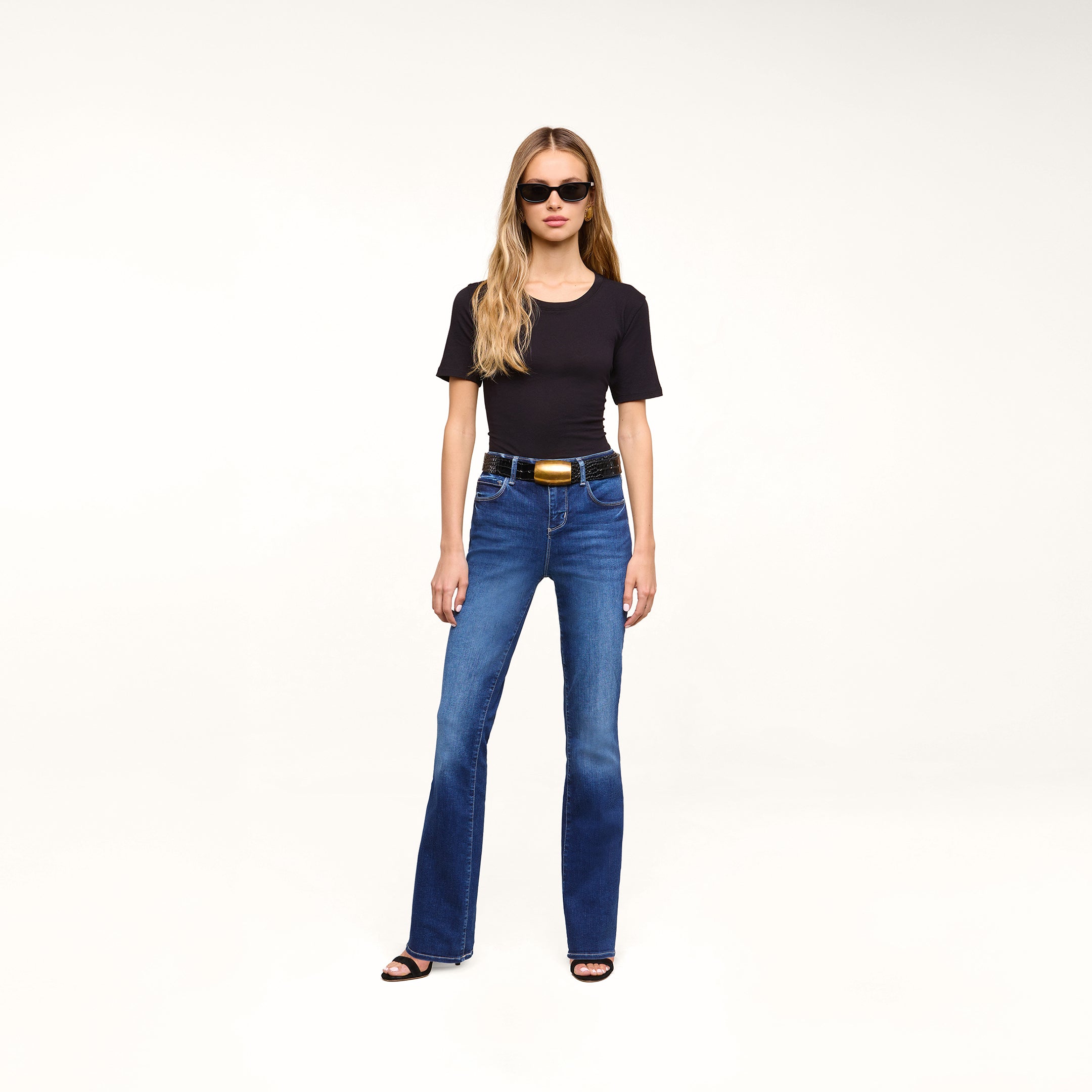 A woman in sunglasses, a black t-shirt, and black heels stands against a plain white background, wearing the Petite Selma Bootcut Jean.