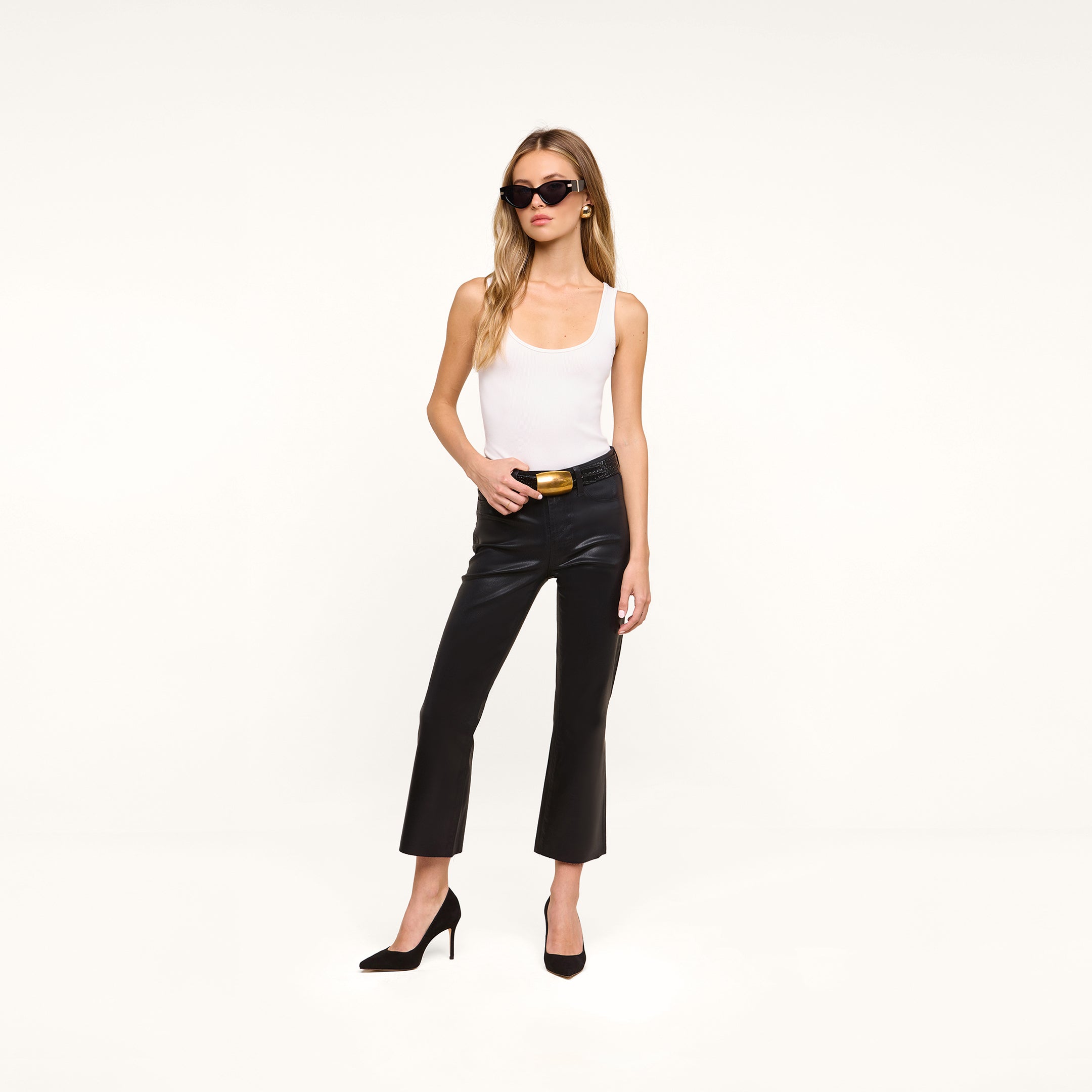 A woman wearing sunglasses, a white tank top, black Petite Kendra Coated Jeans, and black heels stands against a plain white background.