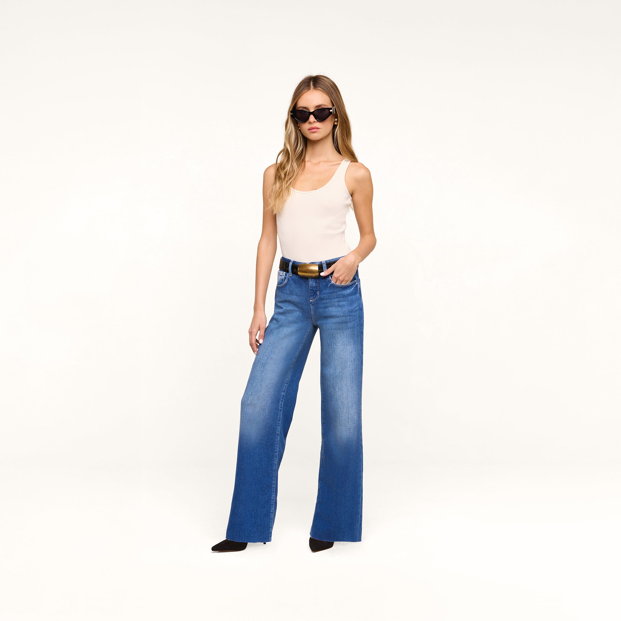 A woman wearing sunglasses, a white tank top, the Petite Scottie Wide-Leg Jean in denim blue, and black shoes stands against a plain white background with one hand in her pocket.