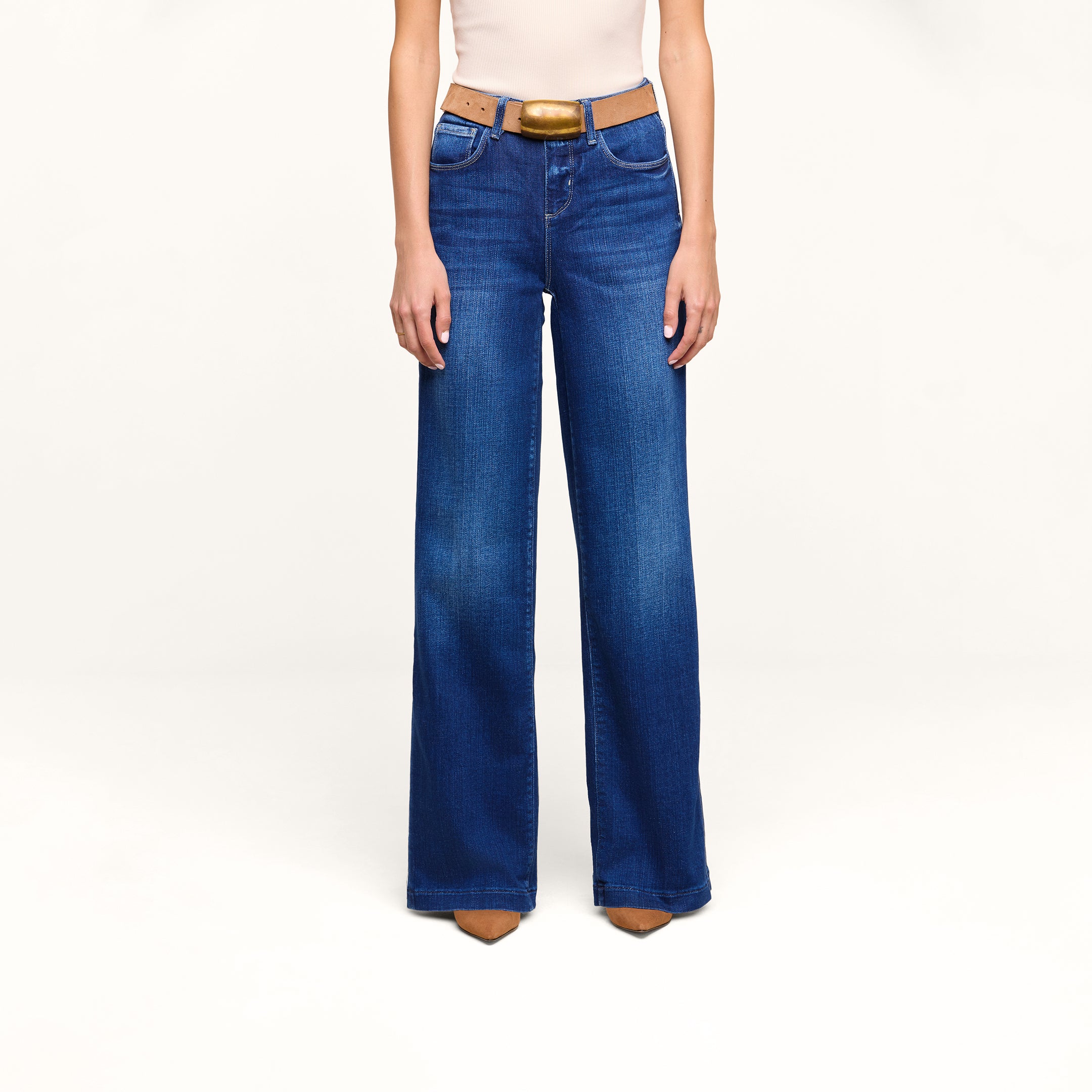 A person stands against a plain white background wearing the Petite Scottie Wide-Leg Jean in blue, paired with a tan belt, beige sleeveless top, and brown pointed shoes.