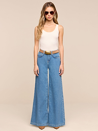 A person wearing sunglasses, a beige sleeveless top, the Petite Quincy Wide-Leg Jean in blue with a brown belt and brown shoes stands against a plain white background.