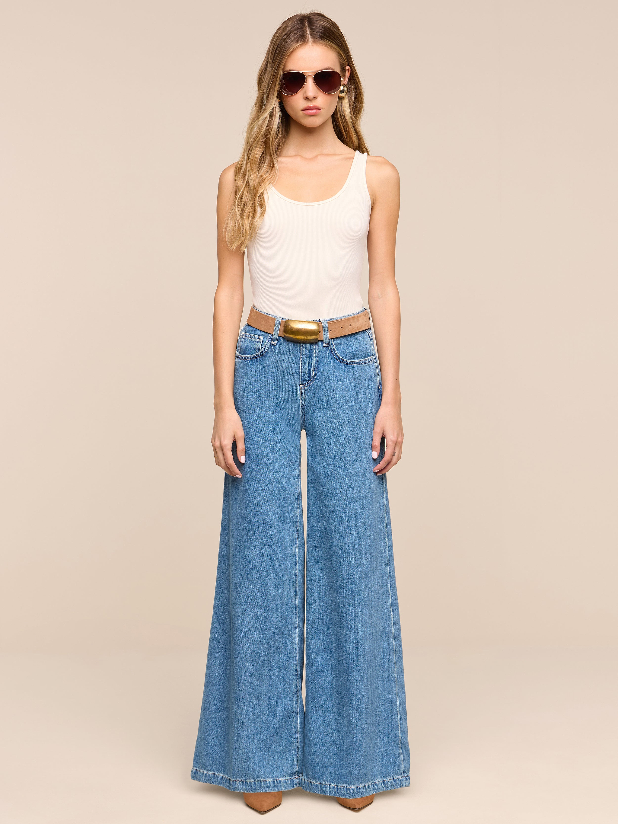 A person wearing sunglasses, a beige sleeveless top, the Petite Quincy Wide-Leg Jean in blue with a brown belt and brown shoes stands against a plain white background.