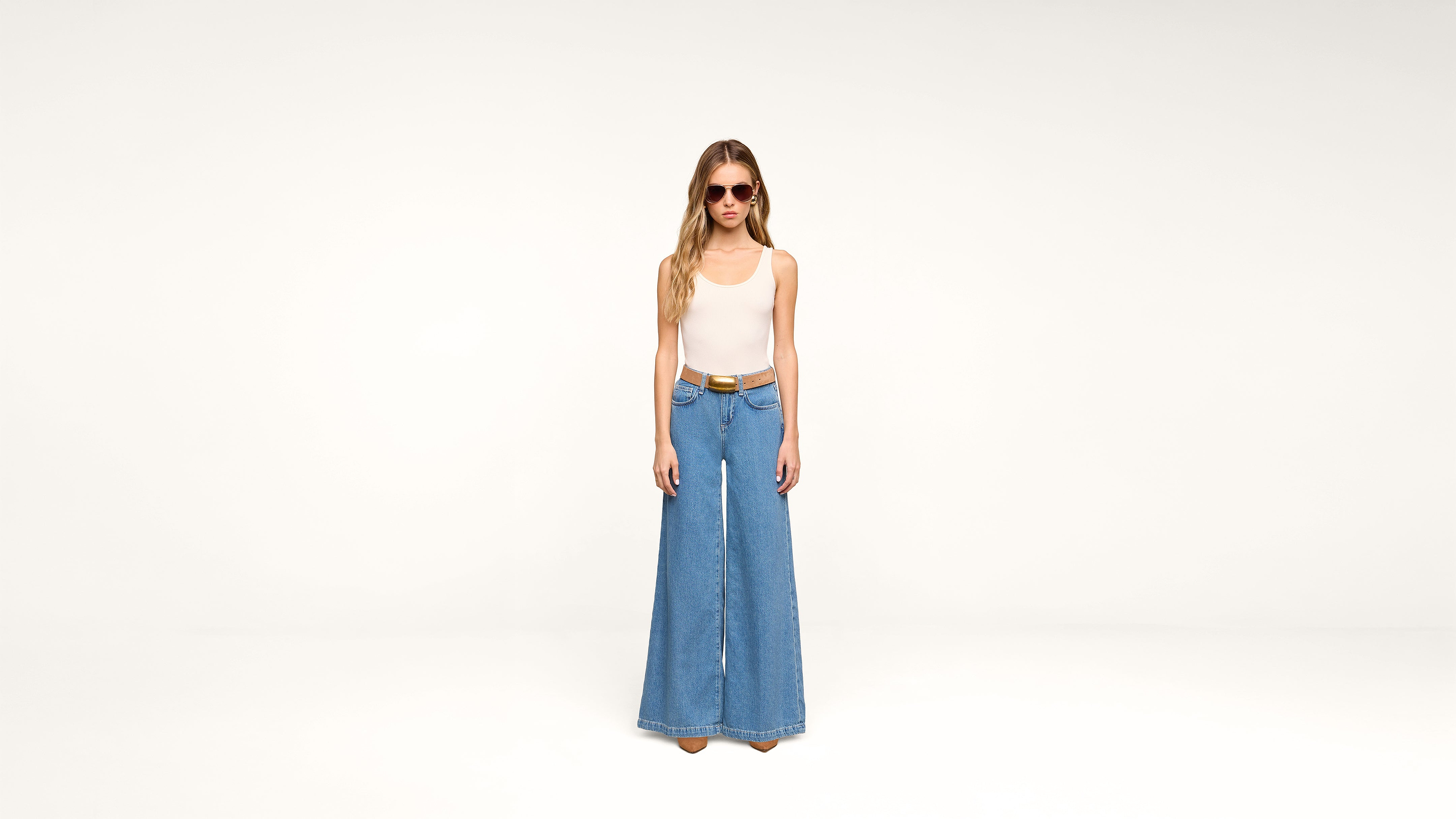 A woman wearing sunglasses, a white sleeveless top, a brown belt, and wide-leg blue jeans stands against a plain white background.