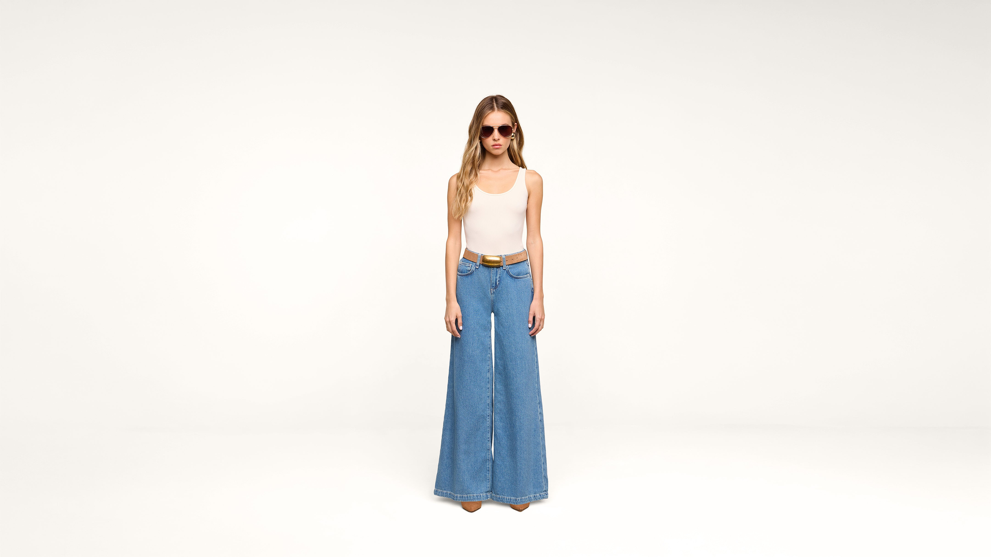 A woman wearing sunglasses, a white sleeveless top, a brown belt, and wide-leg blue jeans stands against a plain white background.