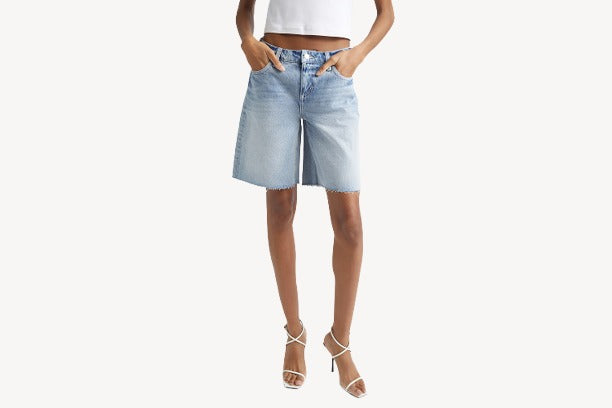 A person wearing light blue knee-length denim shorts, a white sleeveless top, and white strappy heeled sandals stands with hands in pockets against a plain background.