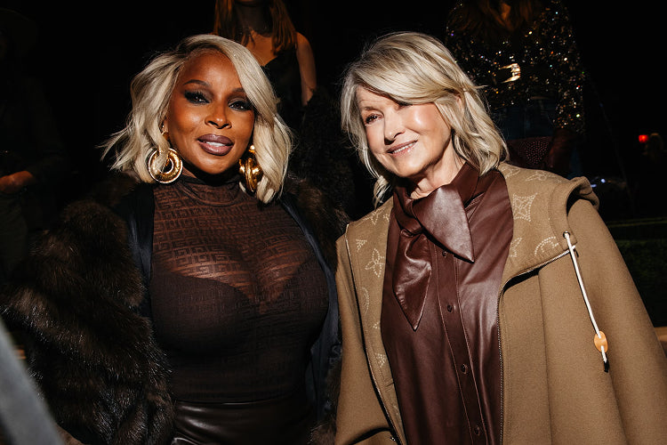 Two women pose together at an event, both wearing stylish brown outfits. One has platinum blonde hair, large hoop earrings, a fur coat, and a sheer top; the other has shoulder-length blonde hair and a tan coat.