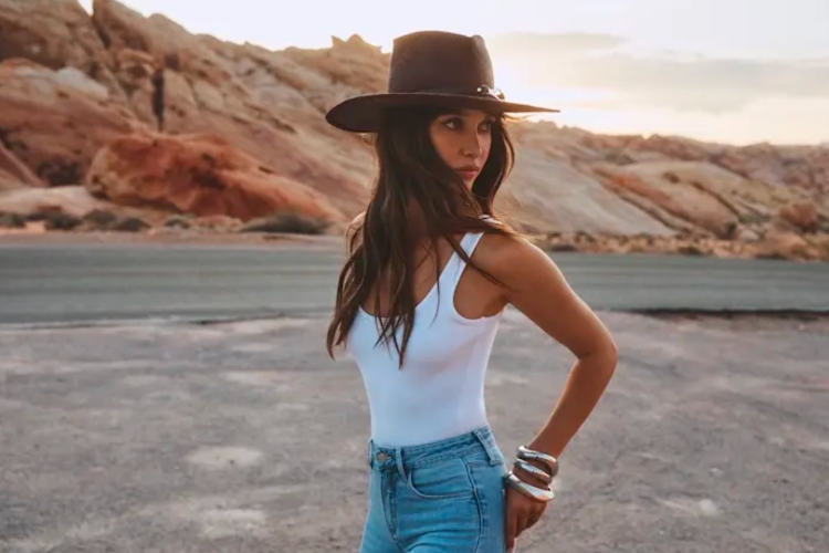 Woman in a white tank top, jeans, and a black hat stands outdoors on a road with rocky desert hills in the background at sunset, looking over her shoulder.