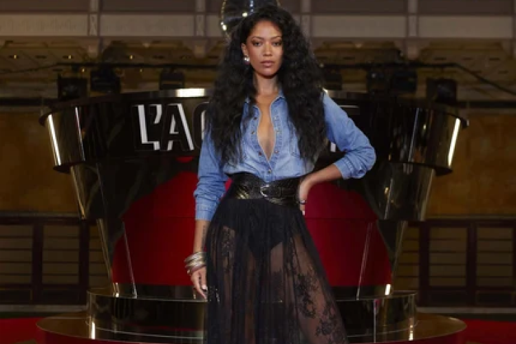 A woman poses confidently in front of a LOréal Paris display, wearing a denim shirt and a sheer black lace skirt with a bold belt, accessorized with bracelets and loose, voluminous hair.