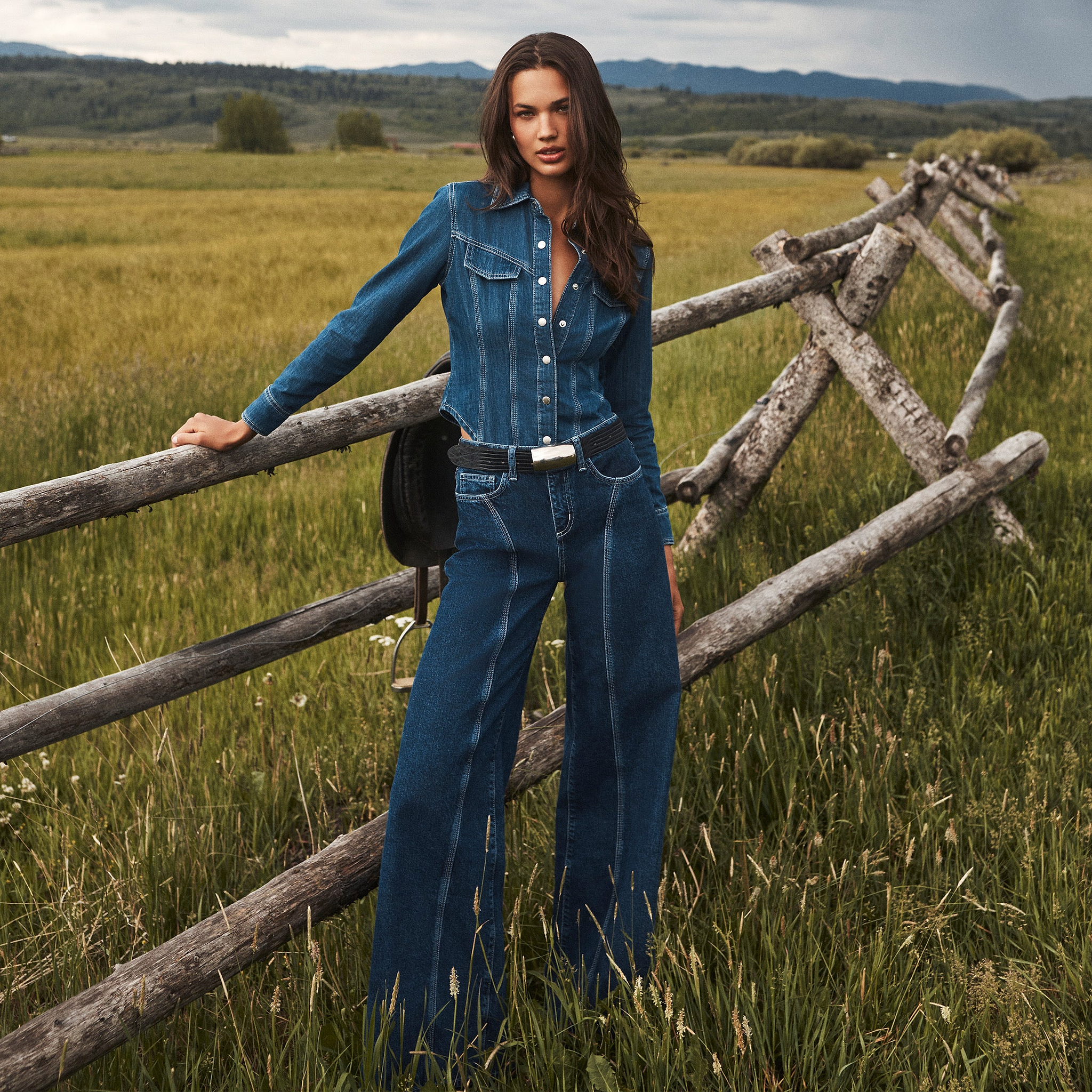 A woman in a denim shirt and wide-leg jeans stands by a wooden fence in a grassy field, with mountains and a cloudy sky behind her.