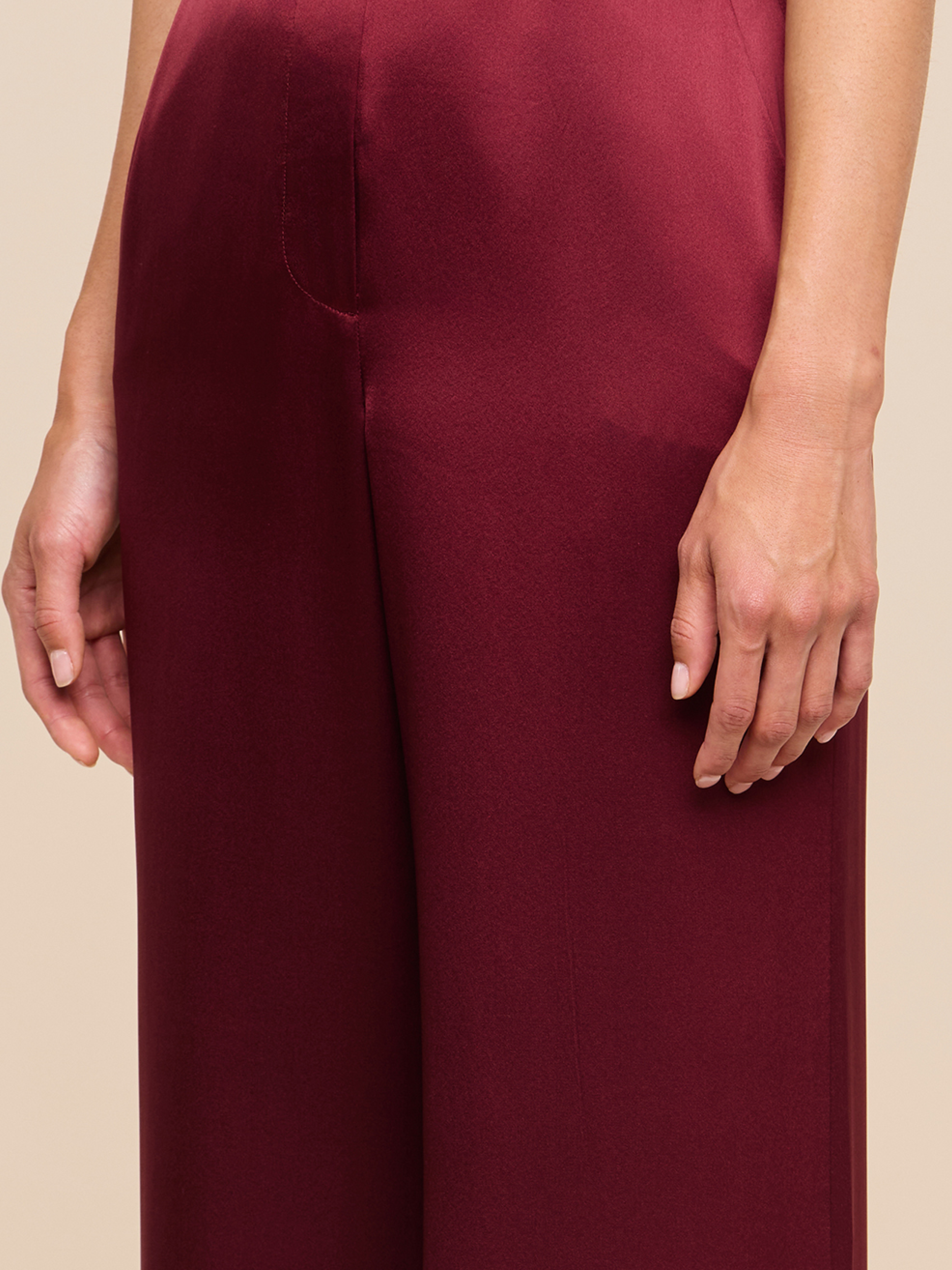 A person wearing high-waisted, burgundy satin pants. Only the lower torso and right arm are visible against a plain beige background.