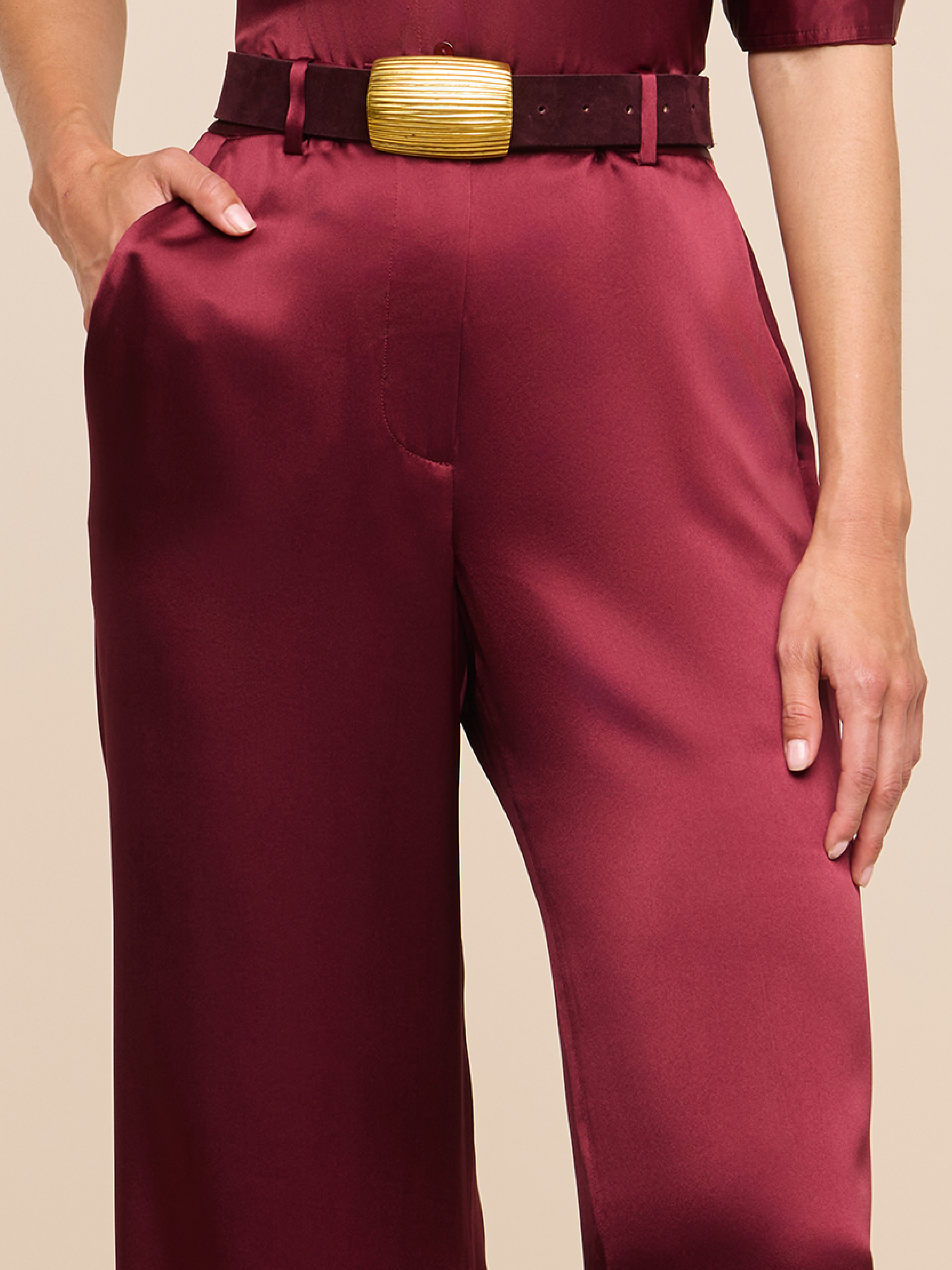 A person wearing burgundy satin pants with a matching top and a wide brown belt with a large gold buckle. One hand is in a pocket and the other rests along the side.