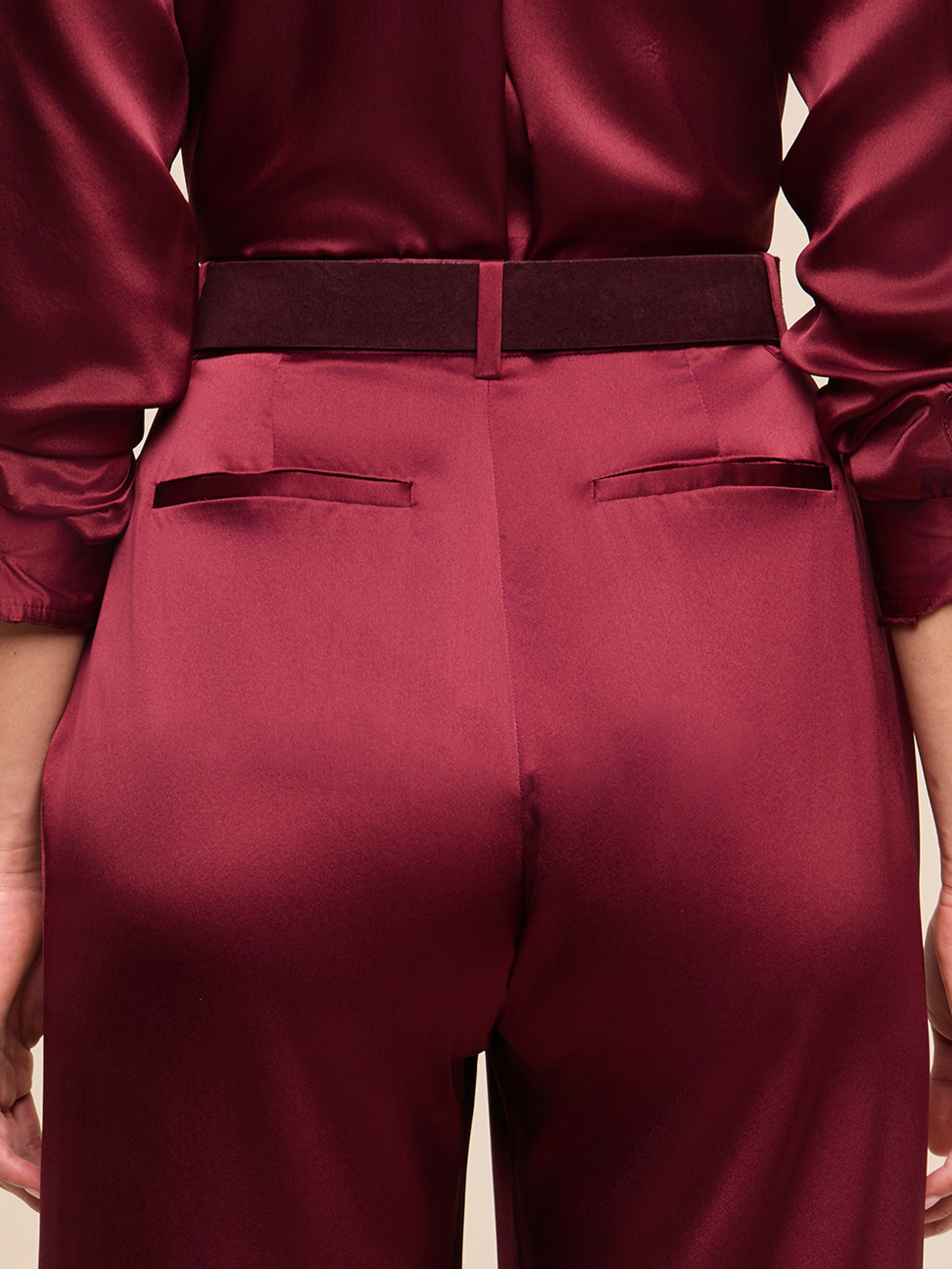 A close-up of a person wearing shiny burgundy satin pants and a matching blouse, viewed from the back, showing the waistband and two back pockets.