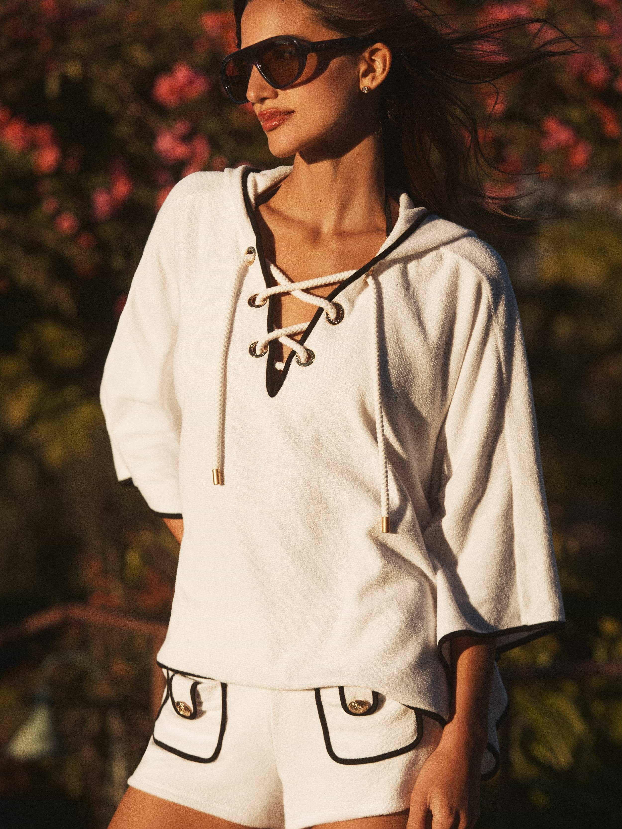 A woman stands outdoors in sunlight, wearing the Demeter Cover-Up Poncho—a white, lace-up terrycloth hoodie with gold accents—paired with matching white shorts with black trim and pockets; blurred flowers fill the background.