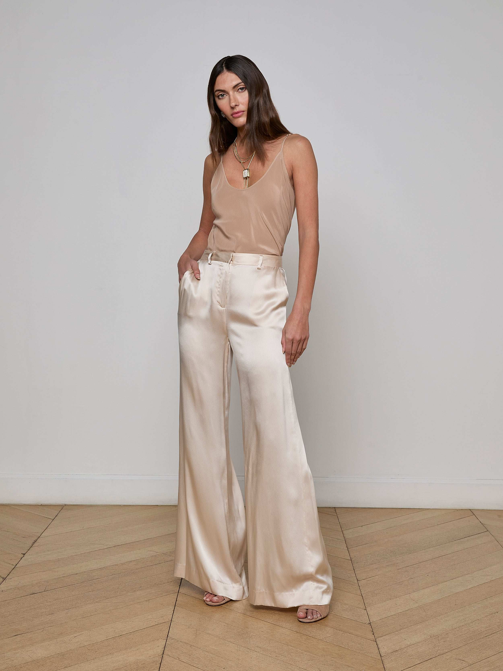 A woman wearing high-waisted, wide-leg Gavin Silk Pants in a subtle champagne hue and a beige sleeveless top stands with her hands in her pockets against a plain white background.