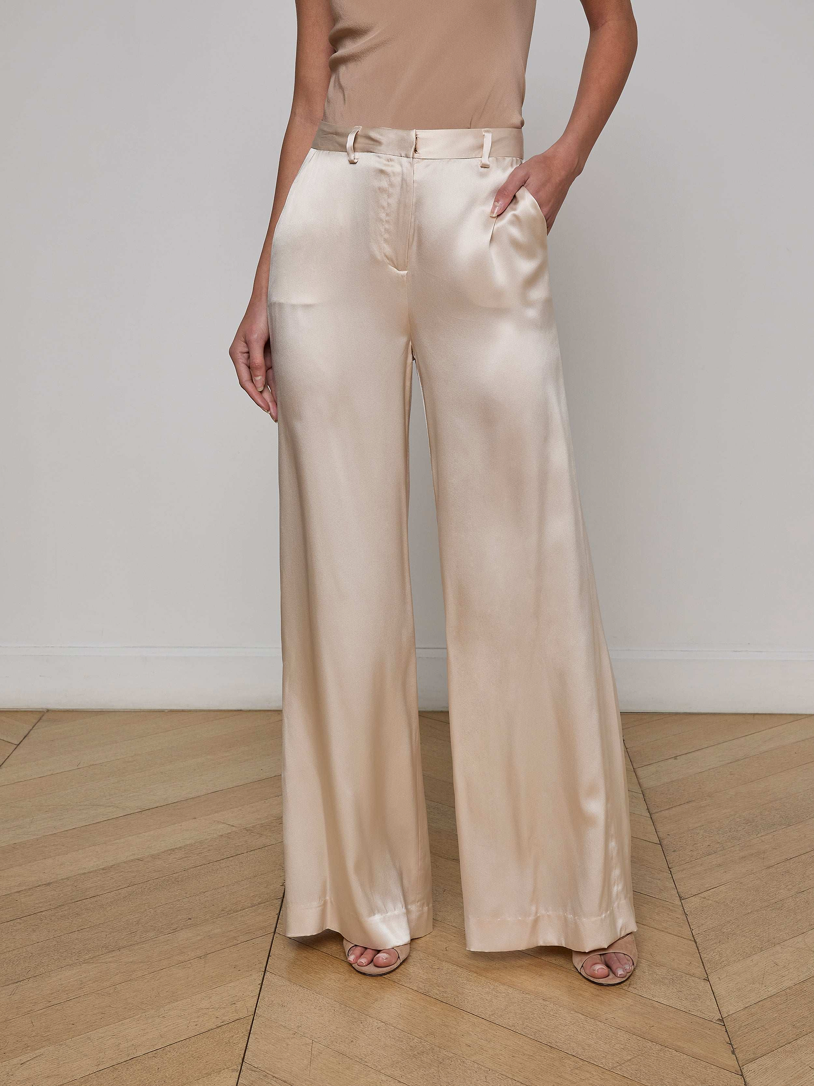 A woman stands against a plain white background, one hand in her pocket, wearing the Gavin Silk Wide-Leg Pant in a light champagne hue paired with a sleeveless top.