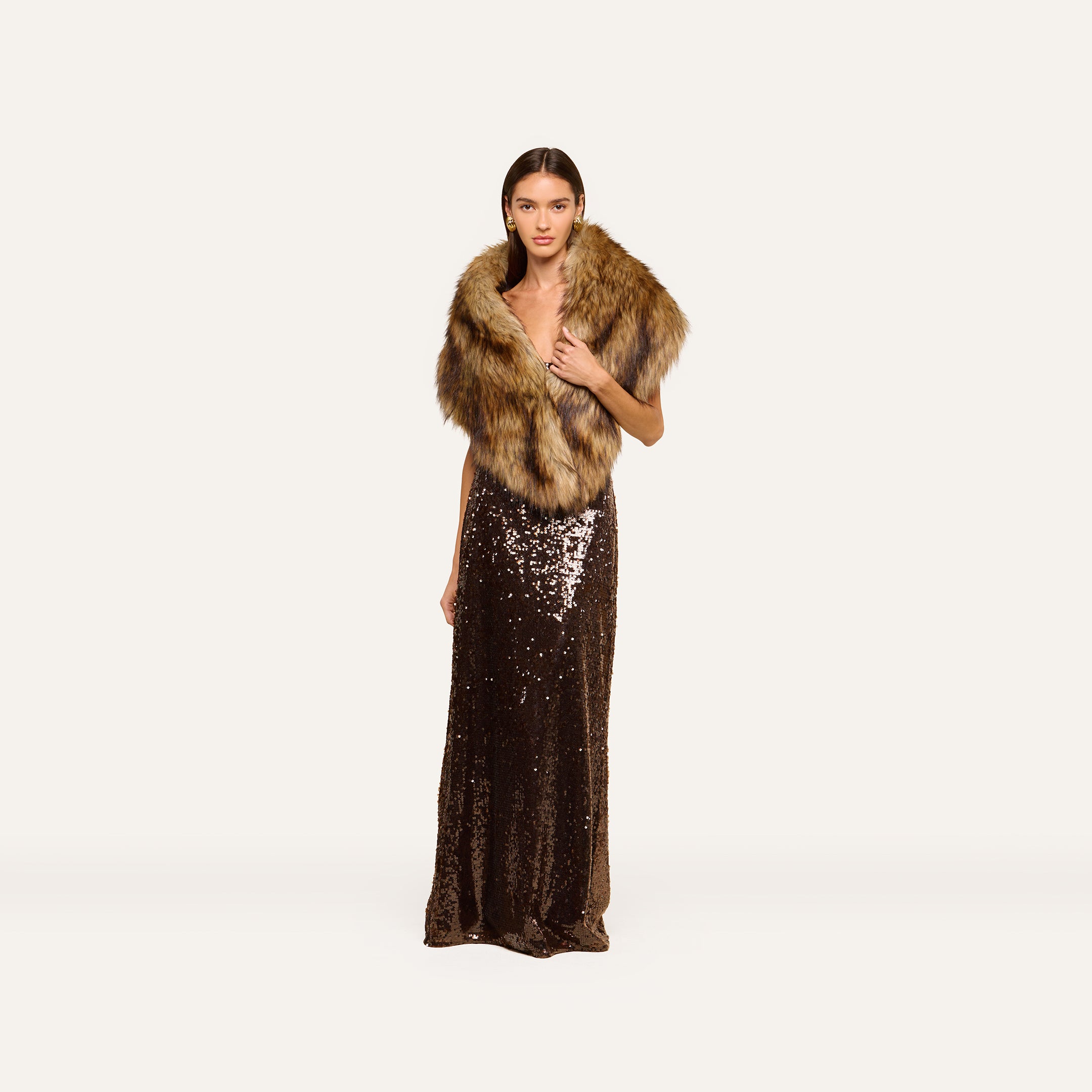 A woman in a long, dark sequin gown exudes plush glamour as she stands against a plain white background, elegantly draped in the Hazel Faux Fur Stole over her shoulders.
