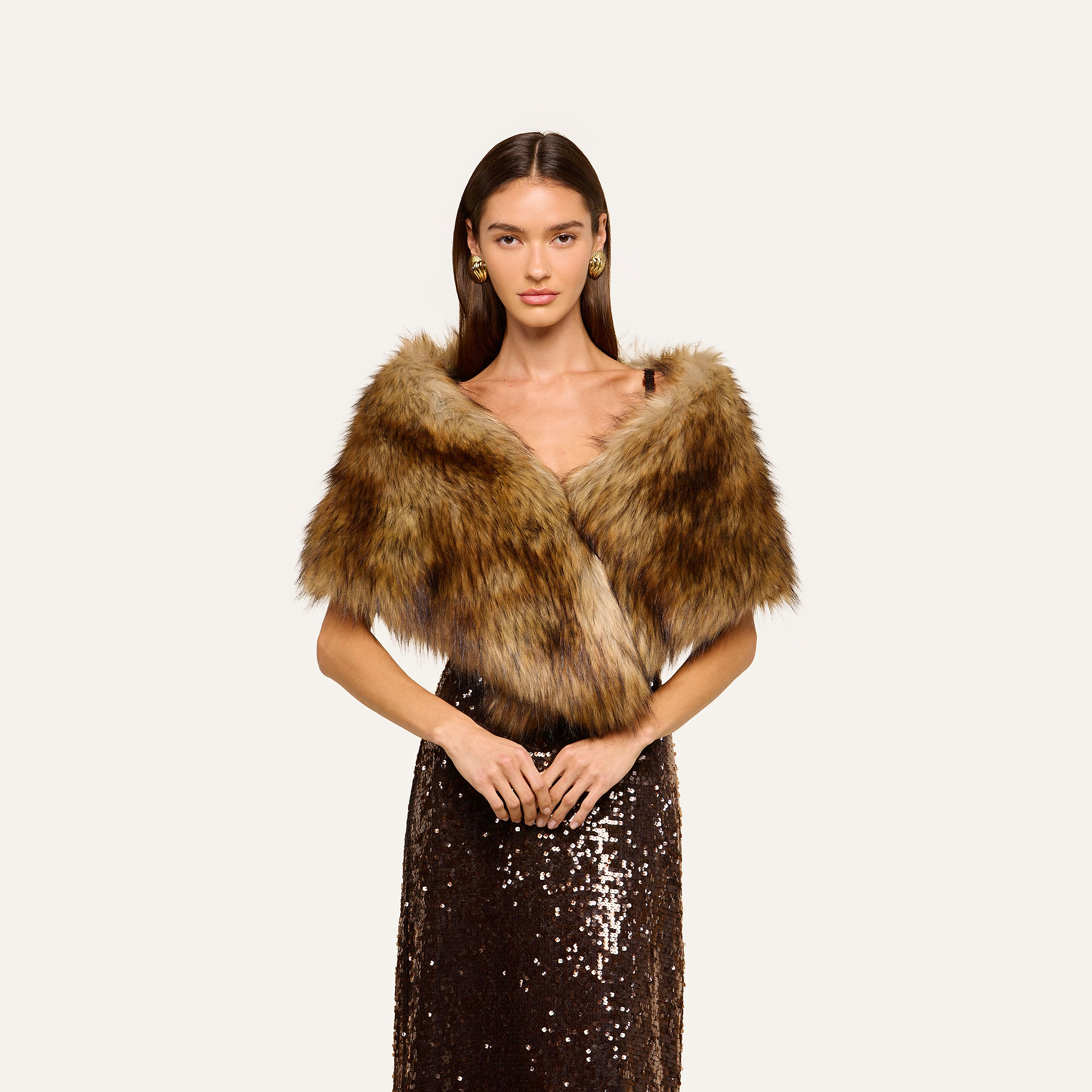 A woman wearing the Hazel Faux Fur Stole in plush glamour brown over a dark sequined dress stands against a plain light background, looking forward with her hands clasped in front of her.