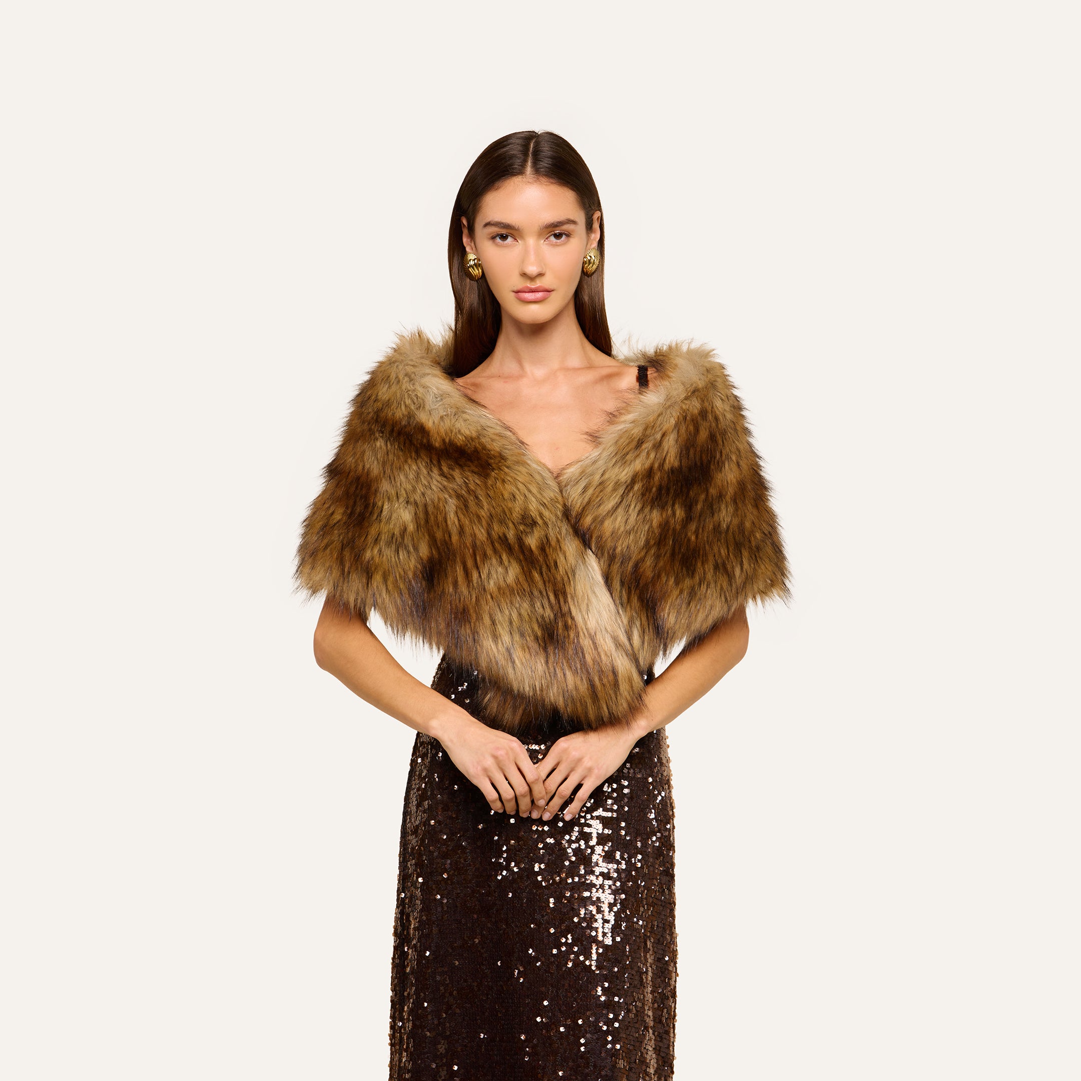 A woman wearing the Hazel Faux Fur Stole in plush glamour brown over a dark sequined dress stands against a plain light background, looking forward with her hands clasped in front of her.