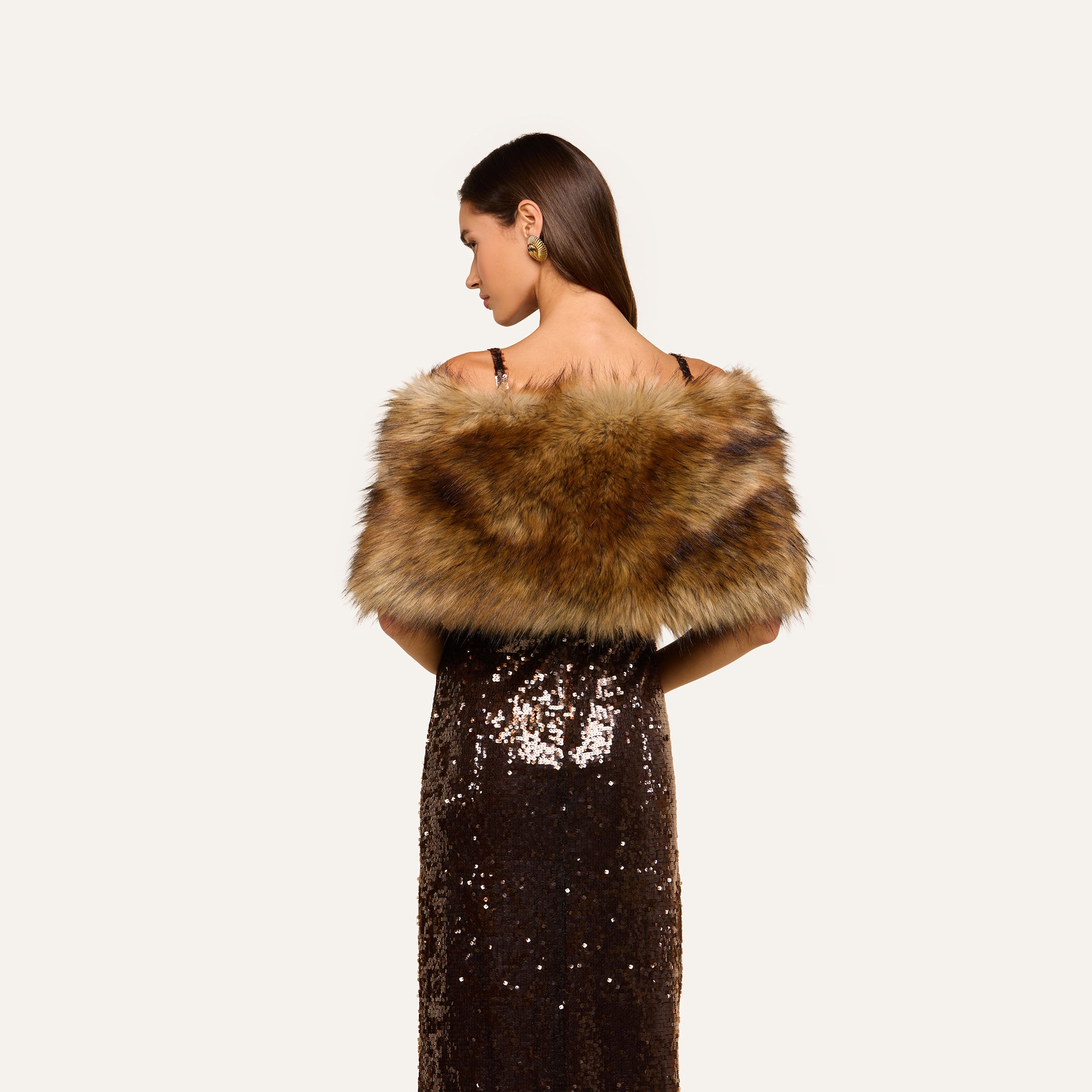 A woman in a dark sequin dress stands facing away, radiating plush glamour with the Hazel Faux Fur Stole draped over her shoulders. Her long, straight hair complements her look, while green earrings serve as her statement accessory against a plain light background.