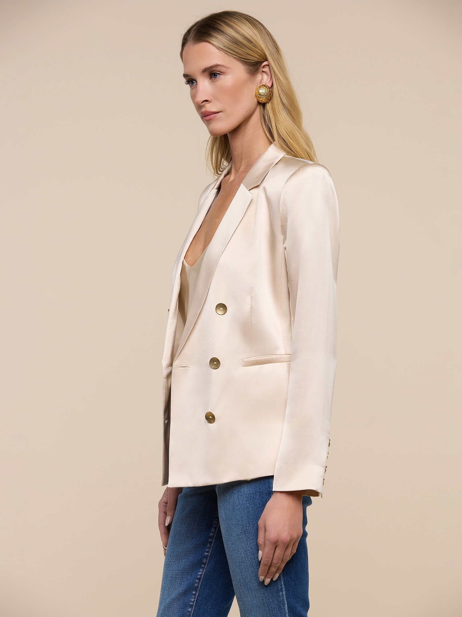 Wearing the Colin Silk Blazer—a double-breasted, cream-colored piece with gold buttons—a woman stands against a plain white background. She pairs the blazer with blue jeans and large gold earrings.