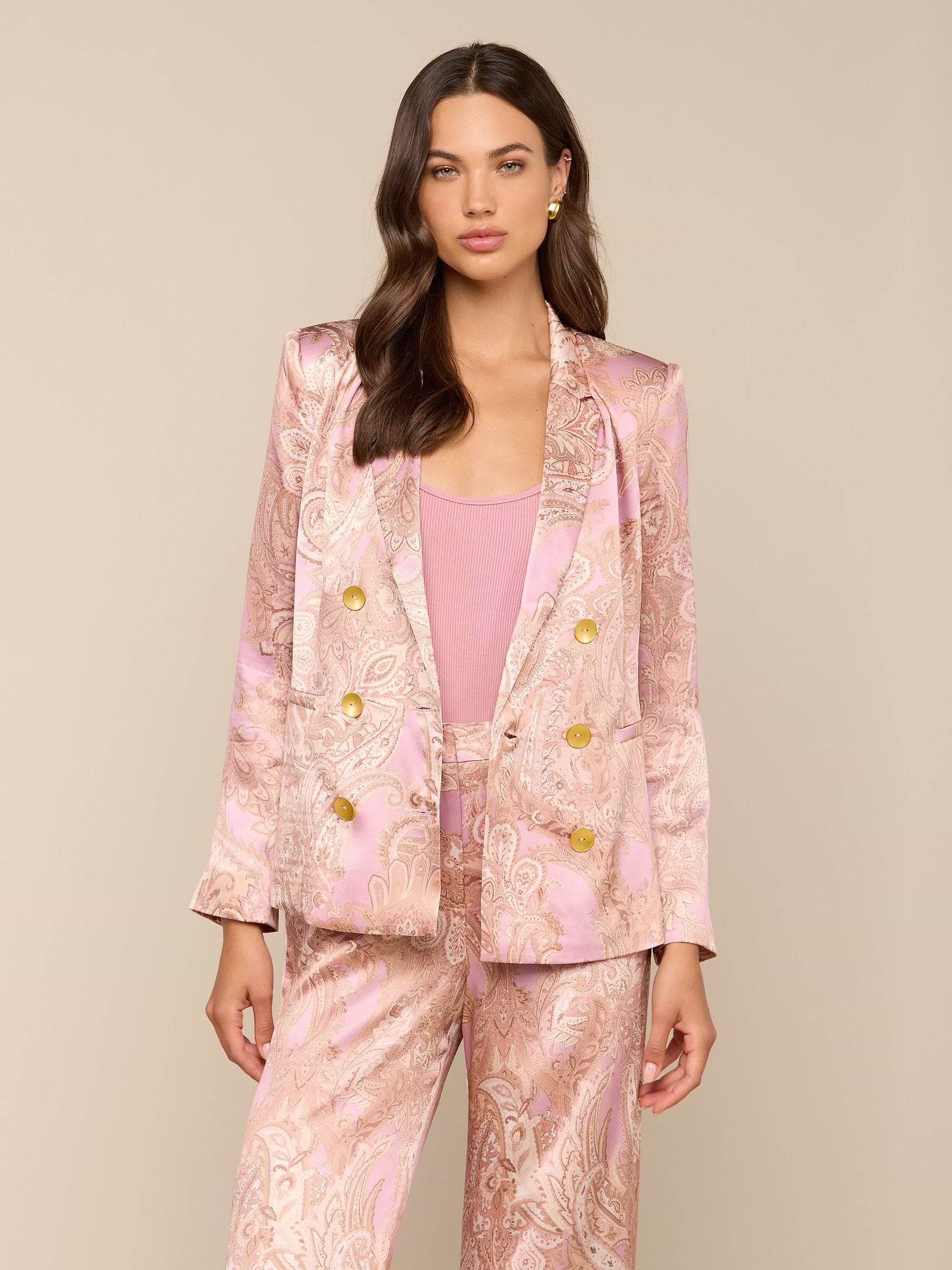 A woman with long brown hair and a neutral expression stands against a plain light background, wearing the Colin Silk Blazer in a pink paisley print with gold buttons.