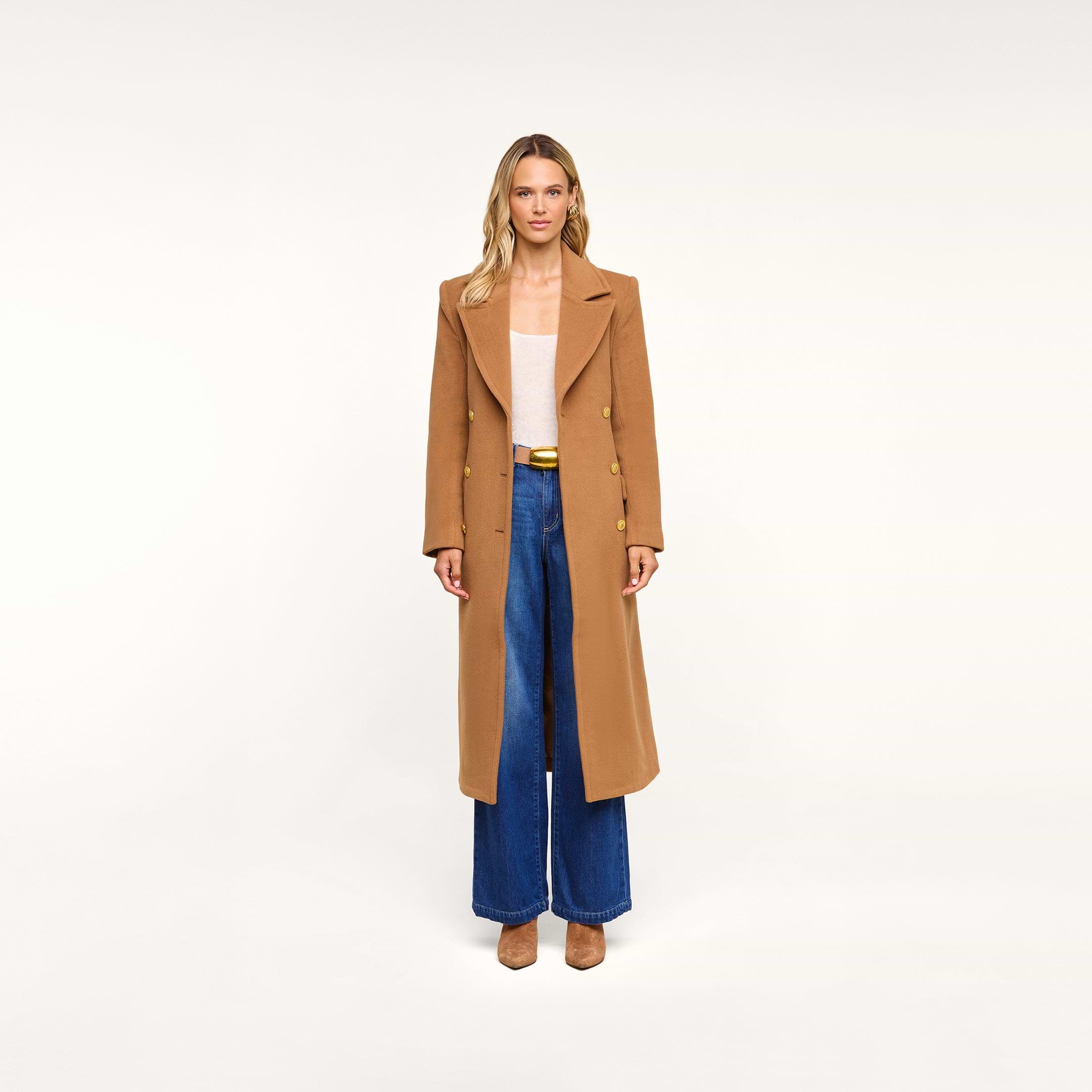 A woman stands against a plain background, facing forward with her arms at her sides, wearing the Kolina Long Coat in Italian wool cognac brown over a light top, paired with blue wide-leg jeans and brown shoes.