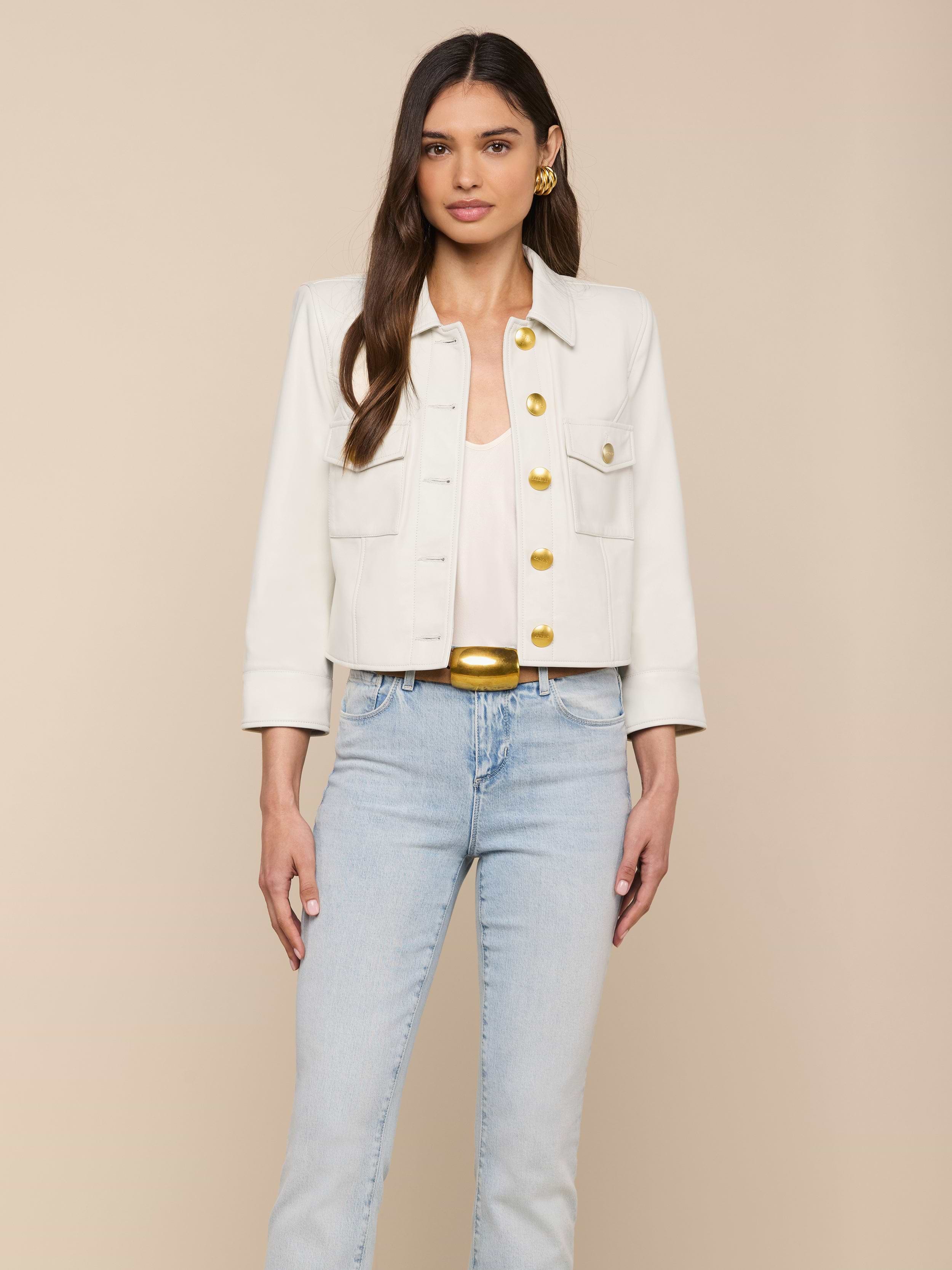 Woman wearing a white Kumi Cropped Leather Jacket with gold buttons, a white top, light blue jeans, and gold earrings, standing against a plain off-white background.