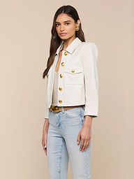 A woman wearing the Kumi Cropped Leather Jacket in white with large gold buttons, paired with light blue jeans and gold earrings, stands against an off-white background.